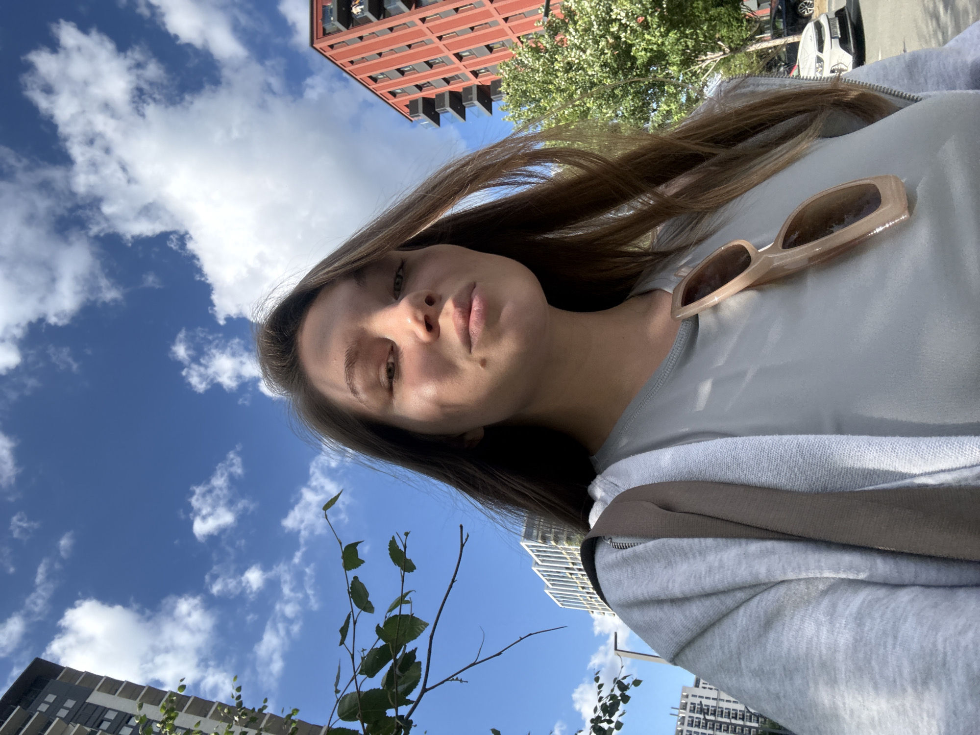 A young woman with long, brown hair, looking at the camera, takes a selfie outdoors on a sunny day. She is wearing a gray t-shirt and a gray zip-up hoodie, with sunglasses clipped to the front of her t-shirt. In the background, tall buildings and trees are visible under a blue sky with white clouds. The location is in the city of Belgrade, Serbia, specifically in front of the buildings of the former "Beko" factory in the Dorćol district, now transformed into a modern residential and commercial complex. The overall scene conveys a casual and urban atmosphere.