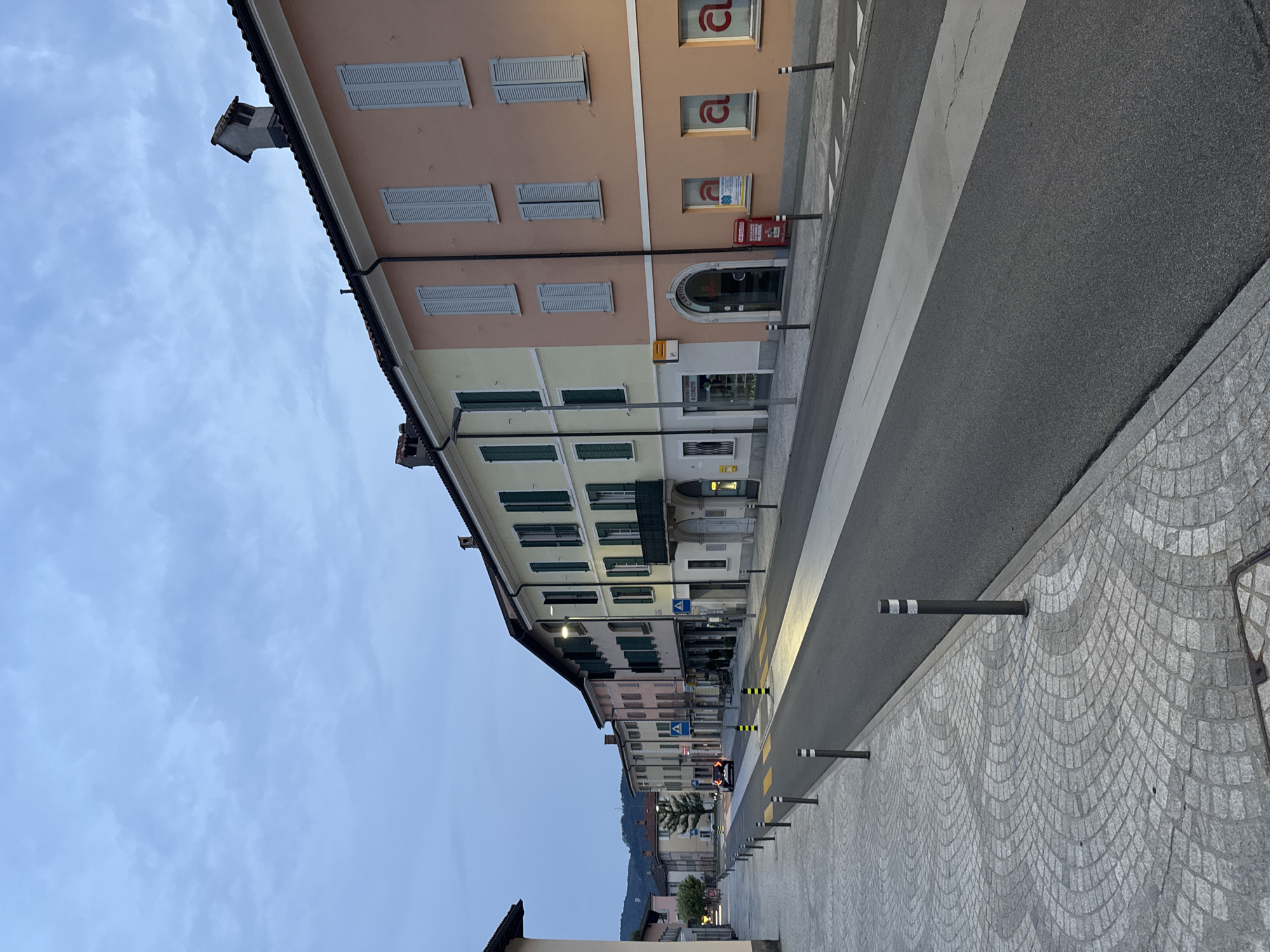The image depicts a street scene in Balerna, Switzerland, likely during dusk or early morning given the twilight sky and illuminated building lights. The perspective is from a pedestrian sidewalk, looking down a gently sloping street lined with multi-story buildings. The buildings are painted in various pastel colors, including pale yellow, pink, and beige, with gray shutters on the windows. Some buildings have architectural details like balconies and chimneys.
The street itself is paved with asphalt and features a pedestrian crossing with white stripes and yellow bollards marking its edges. The sidewalk on the left is paved with decorative gray cobblestones arranged in a curved, fan-like pattern. Black bollards with reflective white bands are spaced along the edge of the sidewalk and street.
In the distance, a single car with its headlights on is visible driving down the street. Further back, trees and more buildings can be seen, with a hint of hills in the background. The sky is overcast with soft gray clouds. The scene suggests a quiet, residential or commercial street in a European town.
