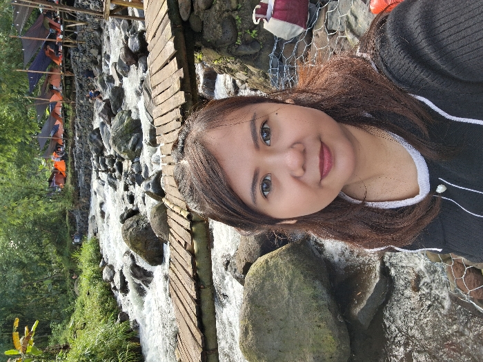 A person poses in a selfie next to a rocky stream. To the left, a wooden plank bridge crosses the water. Camping tents are visible in the background, suggesting a recreational area. The person is wearing a dark-colored top with white trim. The natural environment with the flowing water and vegetation adds to the scenic backdrop.