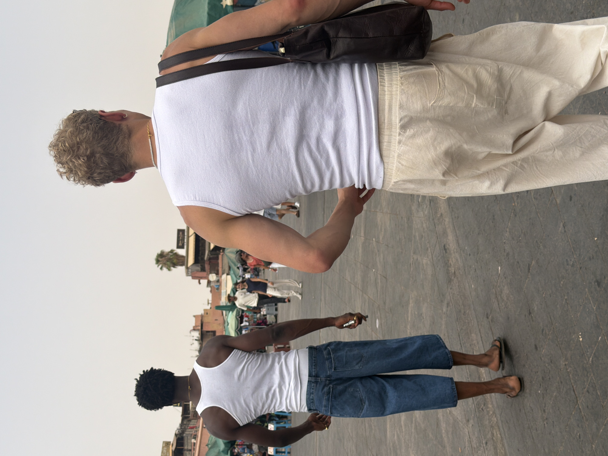 Two men are walking away from the camera on a paved plaza in Marrakesh, Morocco. The man on the left is Black with short, curly hair. He is wearing a white tank top and baggy, blue denim culottes. He has on dark flip-flops and appears to be holding a cigarette in his right hand. The man on the right has blond, curly hair and is wearing a white tank top and loose, cream-colored pants. He has a dark brown leather tote bag slung over his right shoulder. In the background, market stalls with green awnings are visible, and other people are walking in the distance. The sky is overcast.