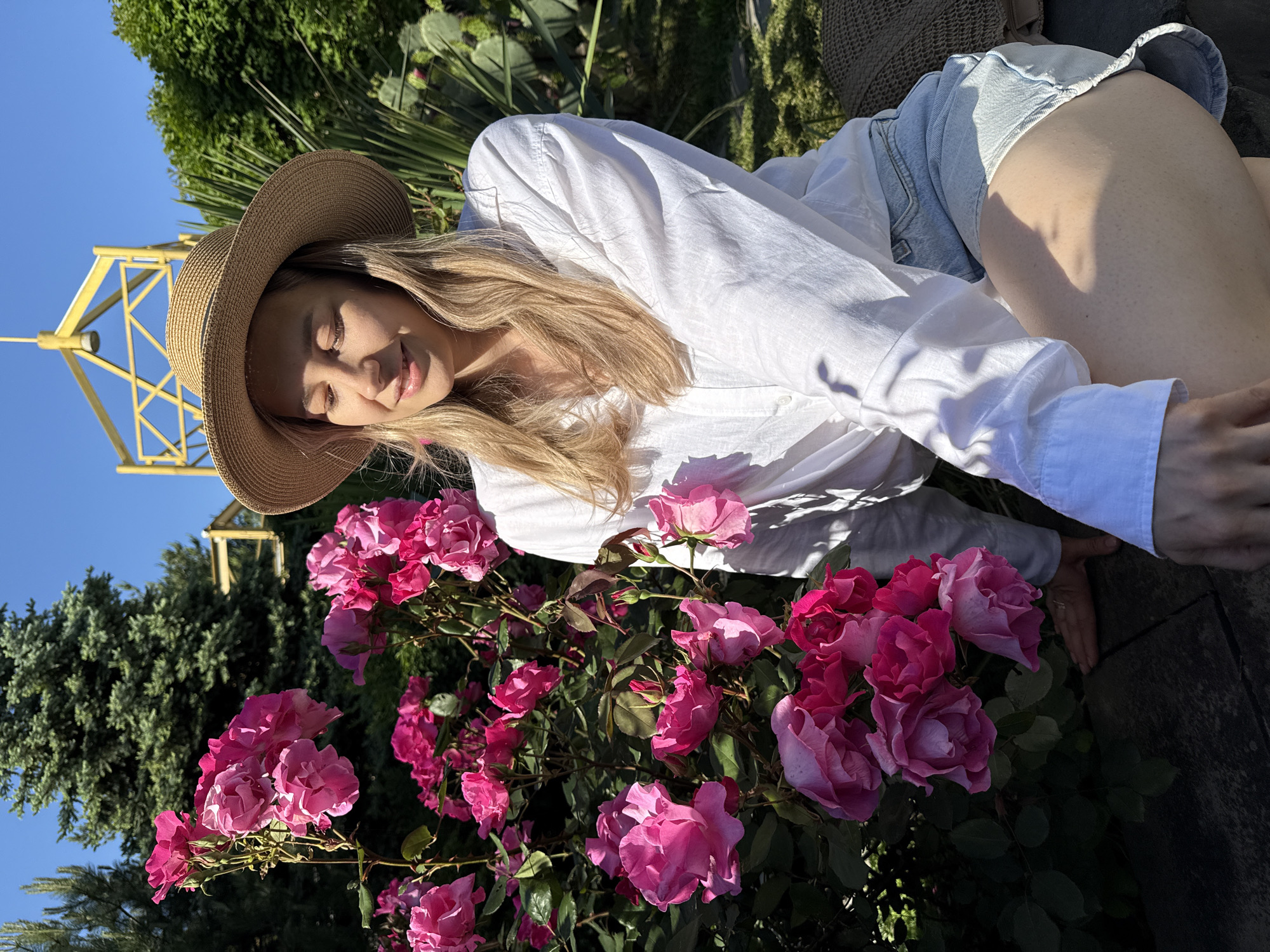 The media file shows a young woman relaxing amongst a vibrant bed of pink roses in an outdoor setting in Nebug, Russia. The woman, with long blonde hair, wears a light straw hat, a white long-sleeved button-down shirt, and light-wash denim shorts. She is positioned as if lying down or leaning into the roses, with her eyes gently closed and a peaceful smile on her face, suggesting a moment of tranquility and enjoyment.
Her hands are near the flowers, with one arm extended and the other bent. The roses are abundant and in various shades of fuchsia and lighter pink, creating a lush foreground. In the background, a clear blue sky dominates, with patches of vibrant green foliage, including coniferous trees and possibly some cacti or succulents, indicating a varied garden environment. A distinct yellow metal structure, possibly an observation tower or artistic piece, is visible against the sky on the left. The scene is brightly lit by natural sunlight, casting gentle shadows and highlighting the textures of the roses and the woman's attire. The overall mood is serene and appreciative of nature's beauty. There is no visible text in the image.