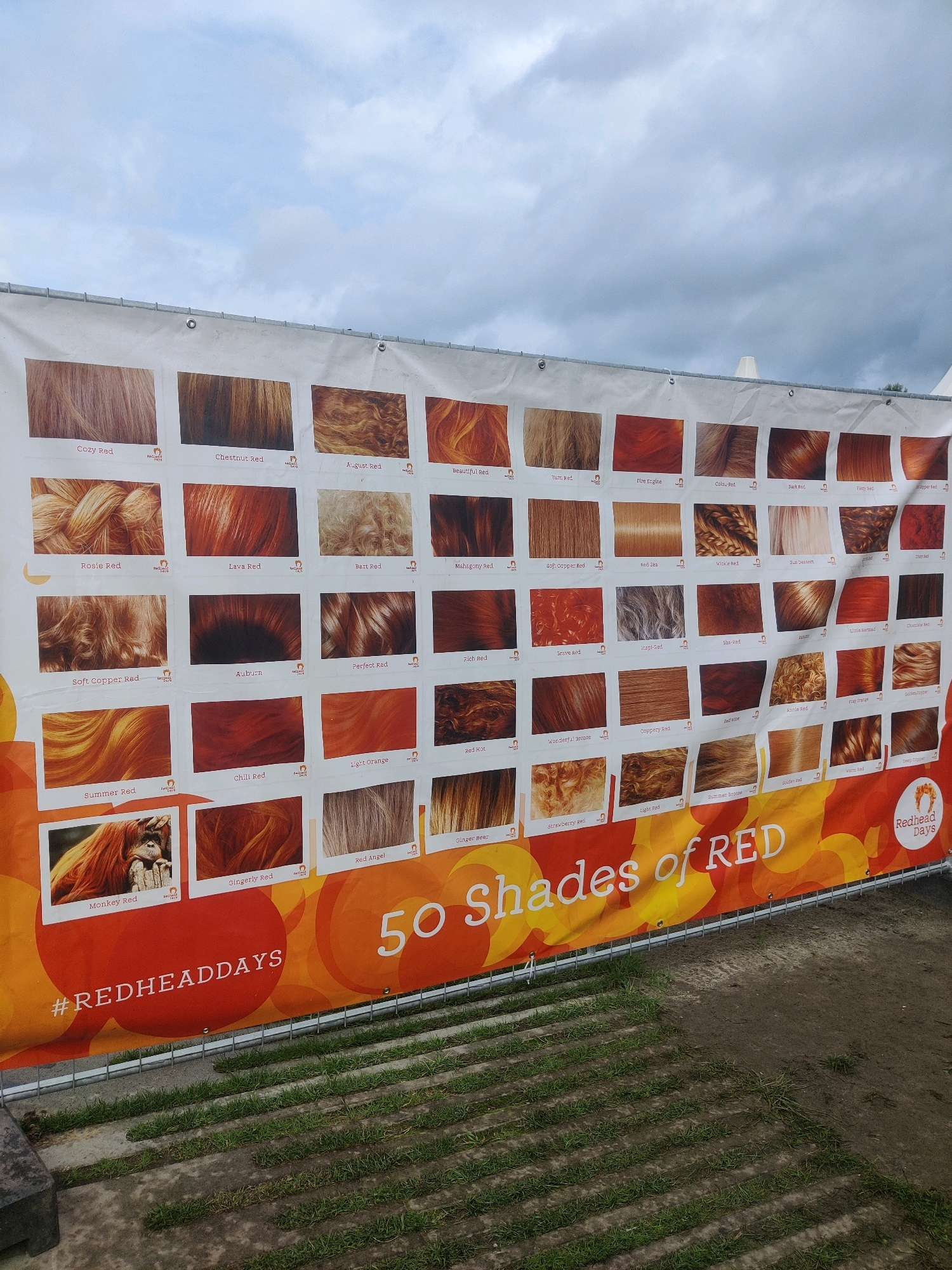 The image shows a large banner displaying "50 Shades of Red" with many swatches of red hair colors. The banner is hung outdoors on a cloudy day, with metal fencing in the foreground. The banner is situated in front of a grassy area with paving stones. The banner is branded with "#REDHEADDAYS" and "Redhead Days", indicating it is from the Redhead Days festival. This festival is held annually in the city of Tilburg, Netherlands. The banner is oriented horizontally and appears to be a prominent feature of the event.