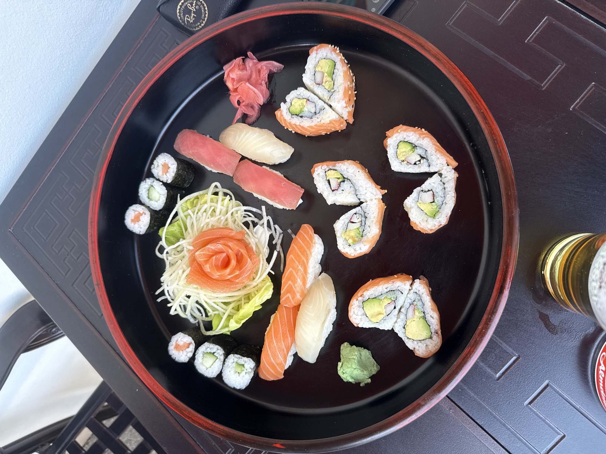 A visually appealing platter of assorted sushi and sashimi is the central focus of this image, presented on a large, round, black serving tray with a distinctive red rim. The tray rests on a dark, possibly wooden, table adorned with intricate, carved or embossed geometric patterns that suggest an Asian-inspired design.
The platter showcases a variety of Japanese delicacies. There are numerous pieces of nigiri, including vibrant orange salmon, deep red tuna, and pale white fish, each served on a bed of rice. Several types of maki rolls are present: smaller rolls containing salmon and cucumber, and larger inside-out rolls (uramaki) filled with avocado and crab stick, some featuring a topping of salmon. Many of these larger rolls are cut in half, revealing their fresh ingredients.
Traditional Japanese condiments are also included: a mound of pink pickled ginger (gari) is placed on the top left of the tray, and a dollop of green wasabi paste sits at the bottom right. A decorative element features thinly sliced salmon sashimi artfully arranged into a rose shape, presented on a bed of shredded white cabbage and a green lettuce leaf.
Beyond the platter, the top left corner of the image shows a black object, possibly a glasses case, with a discernible "Ray-Ban" logo. In the bottom right, a clear glass partially filled with a golden, foamy liquid (likely beer) is visible, next to a red bottle cap with faint, unreadable text. The scene is brightly illuminated by natural light, indicating a daytime setting. This meal is likely being enjoyed in a restaurant or home in Hainburg an der Donau, Austria.