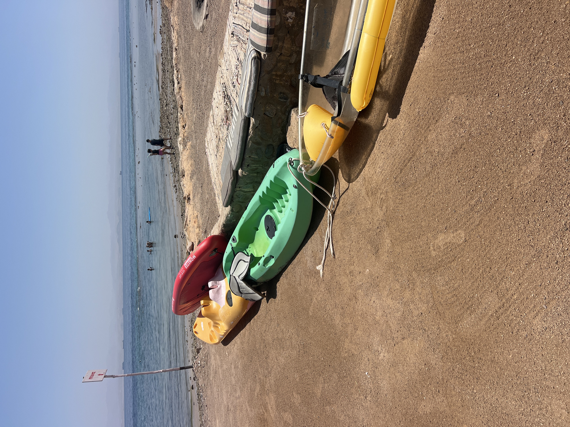 The image depicts a beach scene in an unknown city in Egypt. In the foreground, several kayaks are lined up on the sandy shore. From left to right, there is a red kayak with the word "MONSTER" printed on it, a yellow kayak, a green kayak with a gray seat, a transparent kayak with yellow inflatable pontoons, and another yellow inflatable pontoon. The kayaks are partially on the sand and partially on a rocky area. In the background, the blue sea stretches to the horizon. Several ships are visible on the water. Two people are walking along the shore near the water's edge. Further out in the sea, a few people are swimming or standing in the shallow water. A white pole with a red and white "ENTRANCE" sign is visible on the left side of the image. The sky is clear and blue. The overall environment appears to be a sunny day at the beach, likely a resort or a recreational area.