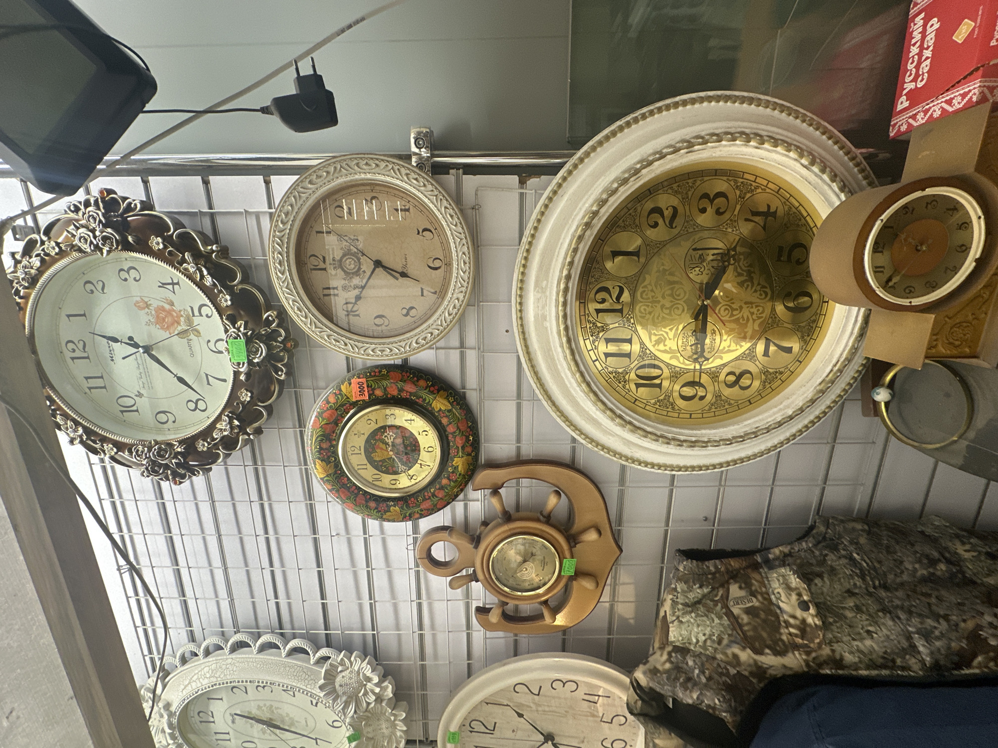 The image depicts a wall display of various clocks in a market or shop. The display is set against a grey grid wall. Prominently featured is a large, ornate, round clock with a decorative brown frame, showing roman numerals and floral motifs on its face. To its left, a white oval clock with a sunflower design is partially visible. Below the ornate clock, a smaller round clock with a folk art design and a price tag is displayed. To the right of the folk art clock, another round clock with a light brown face and roman numerals is mounted. Below these, a wooden clock shaped like an anchor with a central gauge is present. Further down and to the right, a large white and gold round clock with prominent black arabic numerals is displayed. In the foreground, on the left, a camouflage vest is hanging. On the bottom right, a red box with Russian text, likely indicating sugar, is visible. The scene appears to be indoors, with artificial lighting suggested by the reflections on the clock faces. The presence of price tags on some items indicates a retail environment. There is no visible evidence of Novoaltaysk, Russia, in the image itself.