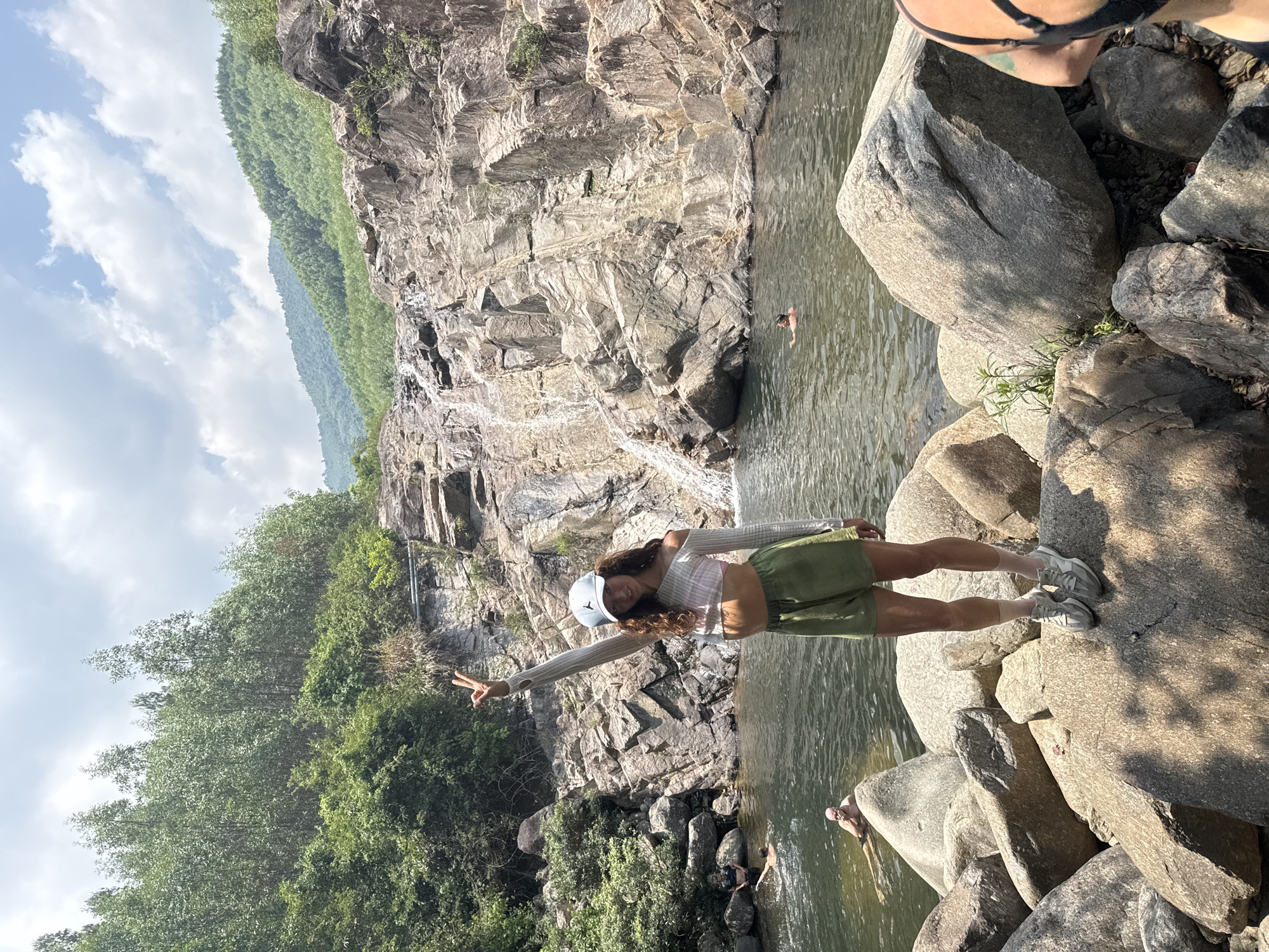 A woman stands on large rocks in a body of water, giving a peace sign. She wears a hat, a long-sleeved shirt, and a green skirt with sneakers. Behind her is a rocky cliff with a small waterfall and green vegetation. Other people are swimming in the water. The location is likely near a waterfall in the mountainous region of Đà Nẵng, Vietnam.