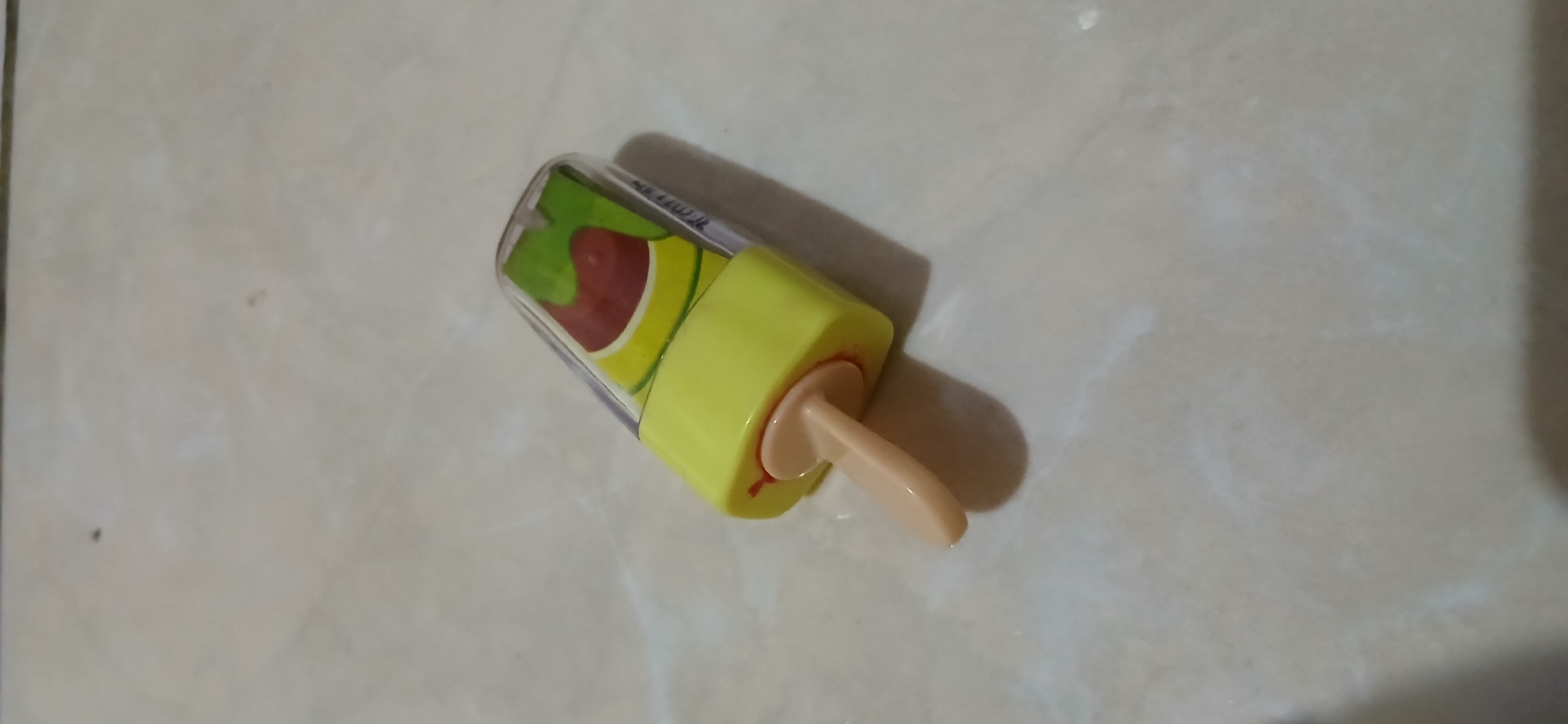 A close-up, high-angle shot captures a novelty ice cream-shaped object resting on a light-colored tiled surface. The object is predominantly yellow with a clear plastic casing at the top, revealing a colorful, illustrated design within, resembling a fruit popsicle. A light brown stick protrudes from the bottom of the yellow base. The tiled floor exhibits subtle variations in color and texture. No people are present, and the scene is devoid of any discernible urban context within Samarinda, Indonesia. The lighting is soft, casting a faint shadow to the right of the object.
