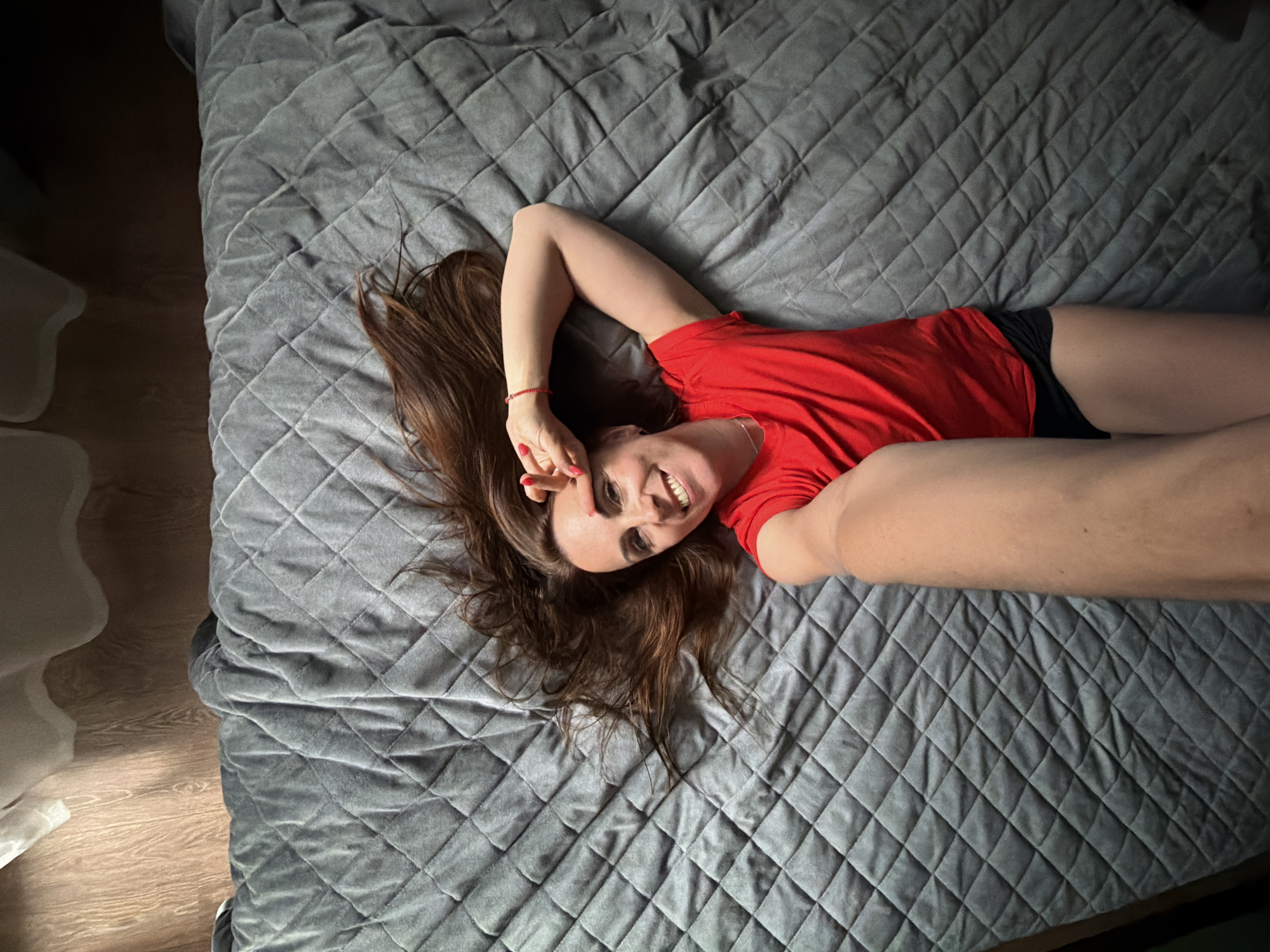 A woman with long, auburn hair is lying on a quilted gray blanket, presumably in a bedroom, as indicated by the wooden floor and what appears to be a curtain in the background. She is wearing a red t-shirt and black shorts. Her right arm is raised behind her head, and her left arm is extended towards the camera, holding what seems to be a smartphone to take a selfie. She is smiling and looking at the camera, with her index and middle fingers forming a peace sign near her forehead. The blanket has a diamond-shaped quilted pattern. The lighting suggests it's daytime, with shadows cast on the floor. The scene is a casual, personal moment captured in a domestic setting. There is no indication of the specific location within the city of Birobidzhan, Russia, as the image contains no identifying landmarks or textual information.