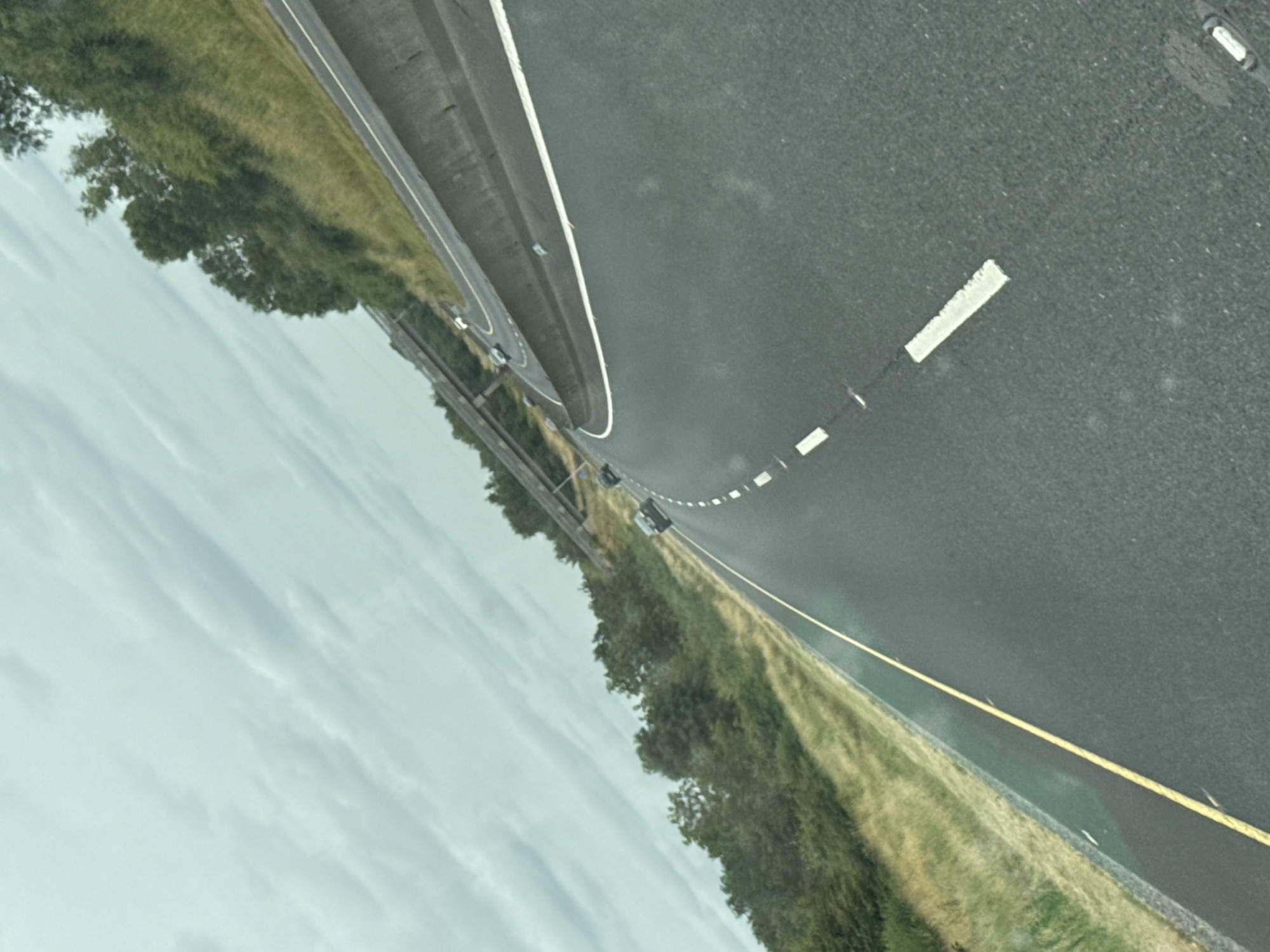 The image depicts a road with a concrete barrier and trees on the side. There are several cars on the road, and the sky is cloudy. The road appears to be a highway, with multiple lanes and a gradual curve. The image seems to have been taken from a moving vehicle, as indicated by the slight blur and the perspective. The overall scene is one of travel on a highway in a rural or suburban area.
