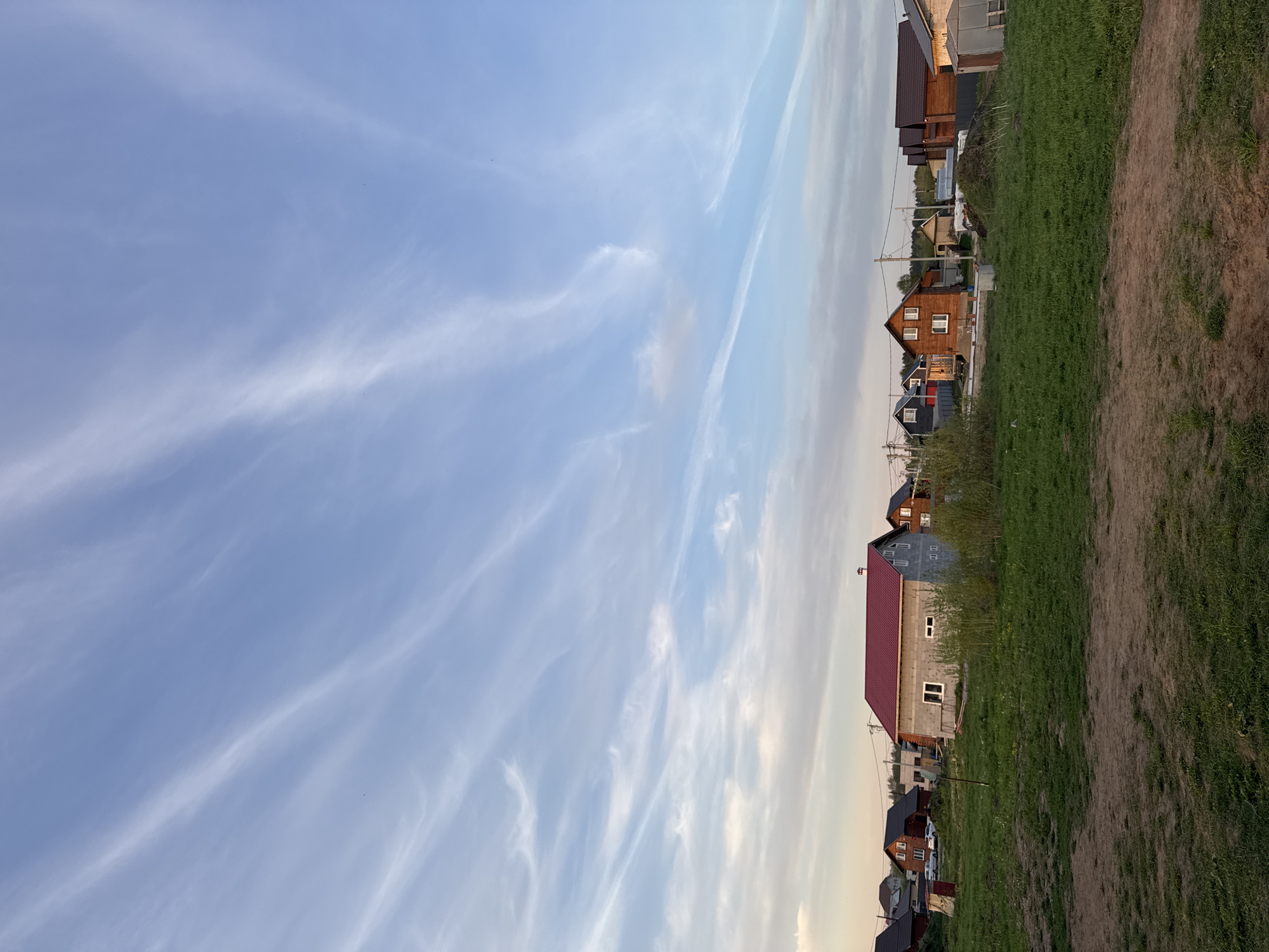 The image shows a neighborhood in Ферма, Russia under a sky with cirrus clouds. Single-family homes line the edge of a grassy and dirt area. Houses are mostly of modern construction, with roofs that are gable or shed type. Exterior finishes are varied, with brick, wood, and light-colored siding. The light suggests the photo was taken near sunrise or sunset. There is no visible human activity in the image.