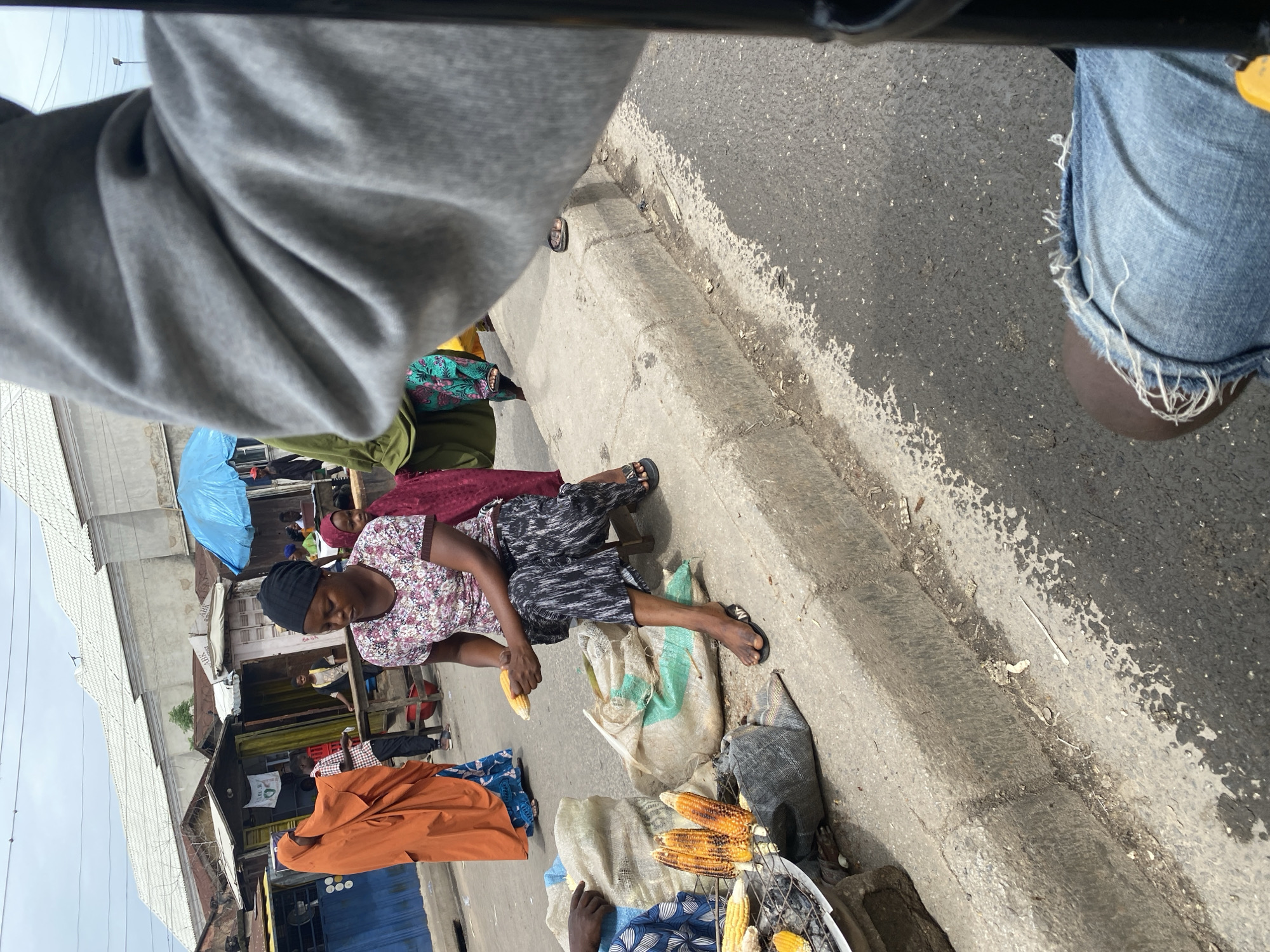 The image depicts a street scene in Jalingo, Nigeria, likely during the day given the bright lighting. The perspective is from someone seated, possibly on a motorcycle or in a vehicle, looking down and to the side.
The primary subject is a woman in the foreground, seated on a low stool beside a pile of corn. She is wearing a floral print blouse, a black headwrap, and patterned pants. Her feet are adorned with black flip-flops. She is holding an ear of corn in her right hand, appearing to be preparing or selling it. Next to her are several bags, one large sack that is partially visible and another lighter-colored bag. A wire rack with several ears of grilled corn sits in front of another person, partially obscured by the bags, suggesting a street food vendor setting.
In the background, several other individuals are visible, some standing and others seated. One person is wearing a bright orange garment, possibly a headscarf or a wrapper. Another person in the far background appears to be wearing a blue shirt. There are stalls or small shops lining the street, with various items, including what looks like fabrics and clothing, displayed. A bright blue umbrella is also visible, providing shade for someone or something behind the vendor. The ground is paved with asphalt, and a concrete curb separates the street from the sidewalk area where the vendors are set up.
The overall atmosphere suggests a busy market or roadside selling area. The subjects appear focused on their activities, with no strong emotional cues evident. The setting is urban and likely a commercial district. No discernible text is visible in the image.