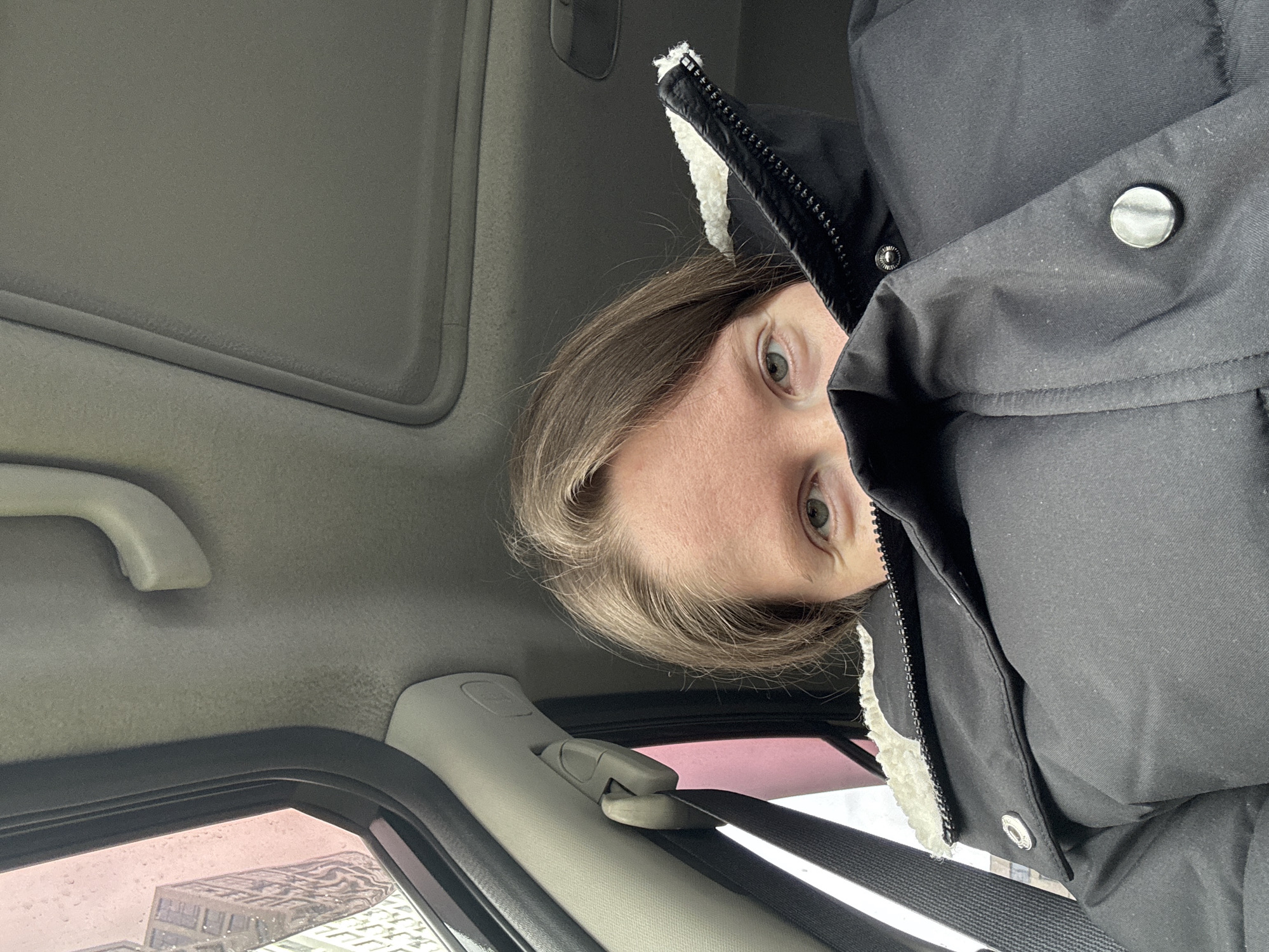 An adult individual is captured from an unusual overhead perspective inside a car. The person's face is partially visible, primarily their wide, light-colored eyes looking directly at the camera, along with their forehead and light brown hair. They are wearing a black winter jacket featuring a white, fluffy lining around the collar or hood, along with metallic snaps and a zipper.
The car's interior details are prominent, including a light grey headliner, a sun visor, a grab handle, and the A-pillar trim. A dark grey seatbelt is visible, buckled into its mechanism, extending across the lower part of the frame. Through the car window, outlines of modern buildings with architectural patterns are discernible under an overcast sky, suggesting it is daytime. The window glass may have slight imperfections or reflections. The scene evokes a casual moment, possibly a selfie, within a vehicle in an urban environment like Khimki, Russia. There is no visible text in the image.
