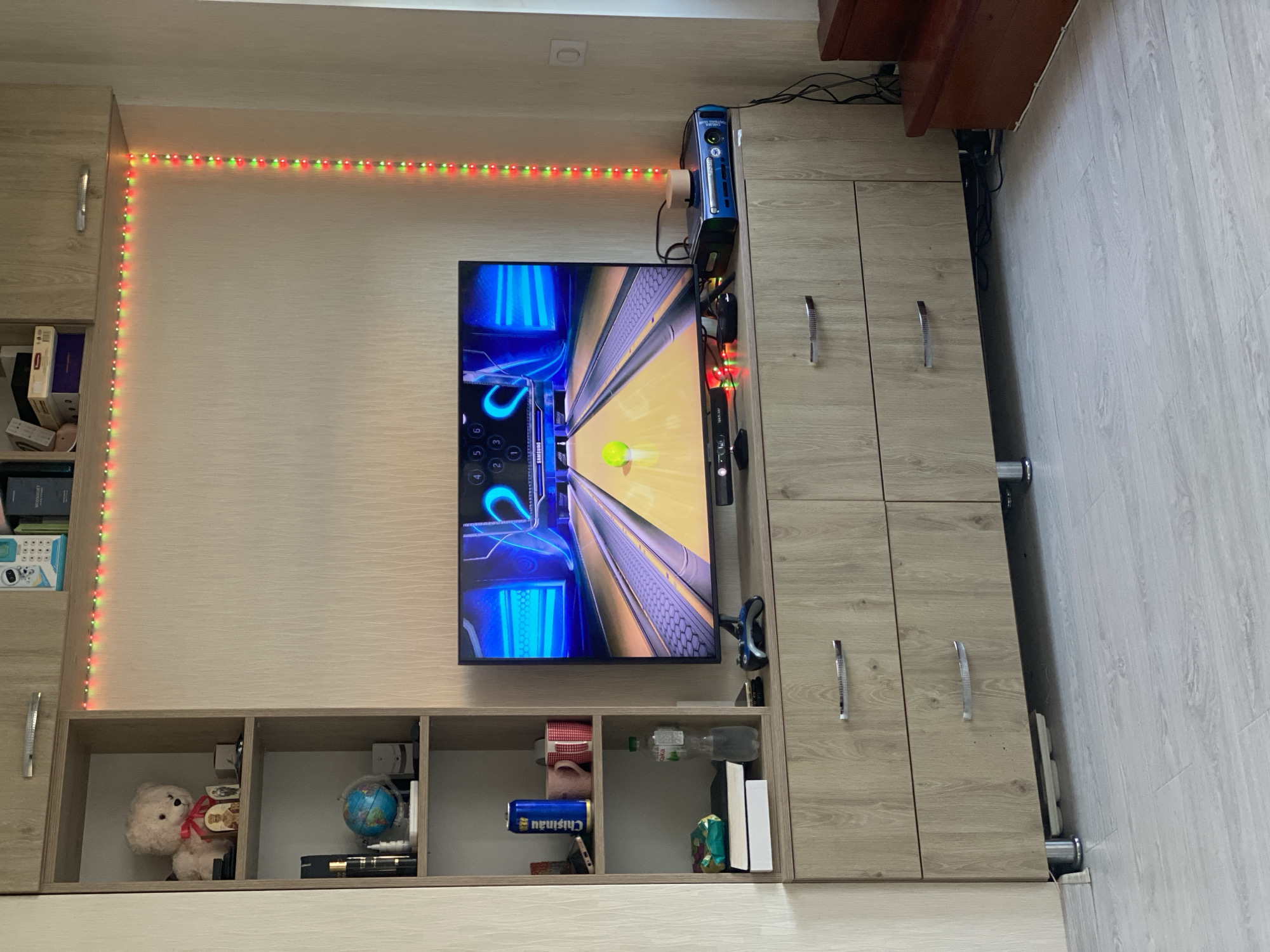 The image shows a room with a TV playing a bowling game, a gaming console, and various shelves filled with items. A light string of red and green lights is outlining the wall above the TV. The room has wooden flooring and cabinets with drawers. There are no people in the image, and no specific landmarks are visible to determine the exact location within Chișinău, Moldova. The scene appears to be a living room or entertainment area.
