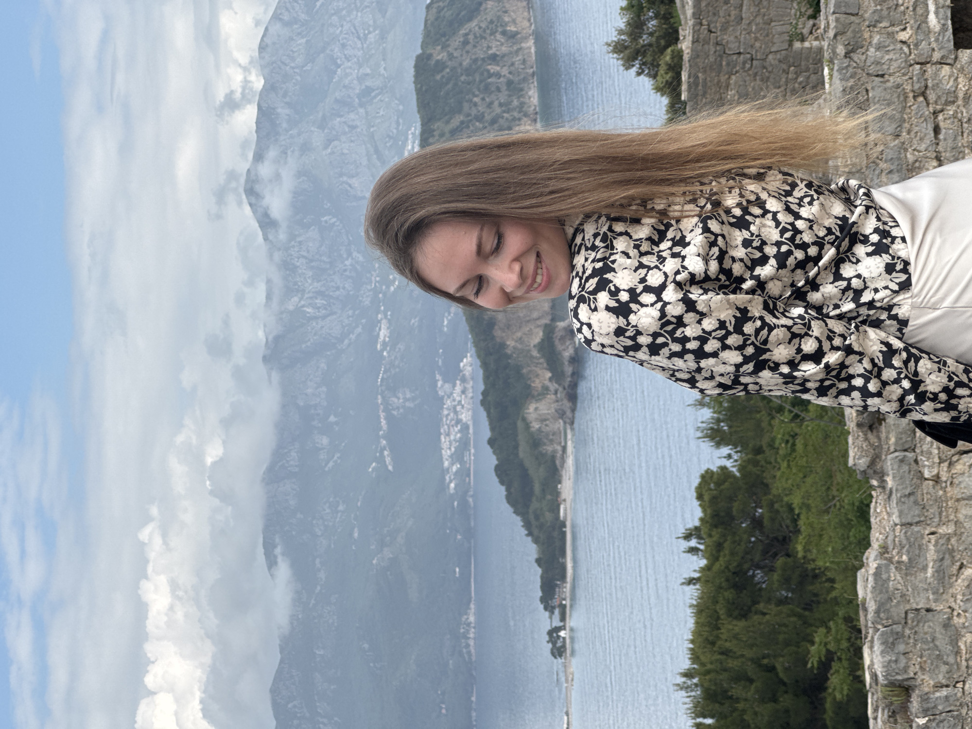 A portrait shot shows a young woman, positioned to the right of the frame, looking down and smiling. She has long, light brown hair that is being blown by the wind. She is wearing a black and white floral shirt and white pants.
The background features a vista of Budva, Montenegro. The Adriatic Sea stretches out, partially obscured by a mountainous landmass with a small town at its base. Further back, a larger mountain range rises, shrouded in clouds.
The woman is standing on a stone structure, presumably a wall or fortification. Vegetation grows below her, towards the lower right corner of the image. The sky is partly cloudy.