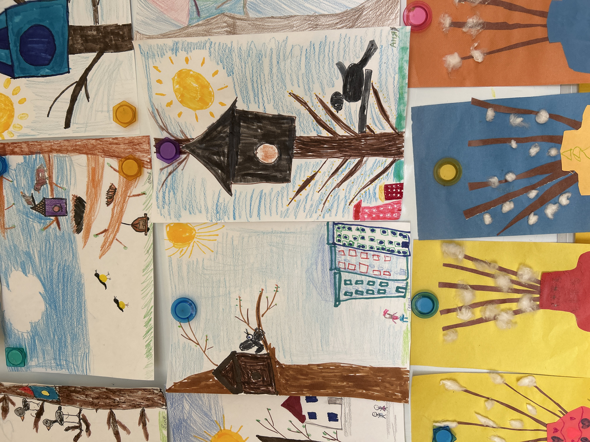 The image shows a collection of children's drawings and crafts displayed on a white surface. The drawings depict various scenes, including birdhouses with birds, suns, buildings, and what appears to be pussy willow branches. The drawings are made with crayons and markers on white paper. The crafts involve paper cutouts and cotton balls to simulate pussy willows. The drawings are held in place by colorful bottle caps acting as magnets. The artwork exhibits a naive and child-like style. The location is likely an educational setting, such as a classroom or daycare. The specific location in Moscow, Russia is not discernible from the image.