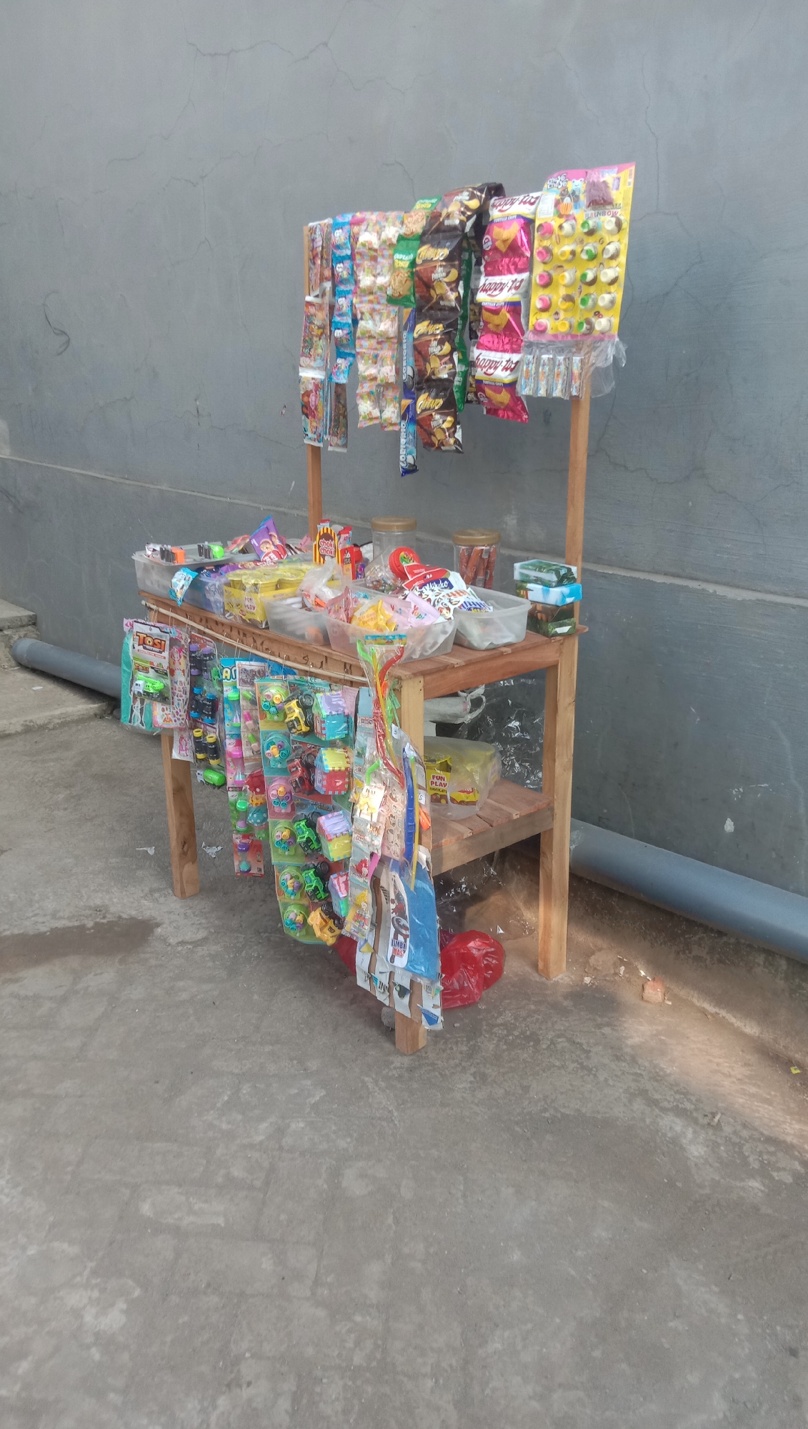 A small, wooden stand displaying an assortment of snacks and small toys is positioned against a textured gray wall. The stand has two shelves. The top shelf holds various packaged snacks, including colorful bags of chips and candies. Some items are in clear plastic containers, while others are displayed in small boxes. The lower shelf features more packaged snacks and several rows of small toys, such as miniature cars and other trinkets, hanging on hooks. The toys are brightly colored and come in blister packs. A red plastic bag rests on the ground beneath the lower shelf. The ground in front of the stand is paved concrete. A gray pipe runs horizontally along the lower left side of the image, near the wall. The scene appears to be an outdoor street stall, likely located in a residential or commercial area of Karang Anyar, Indonesia, given the typical appearance of such vendors. There are no people visible in the image.