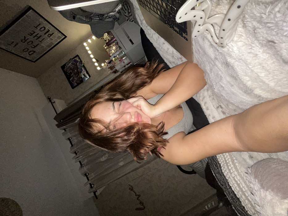 A young woman with shoulder-length brown hair is lying on a bed with a fluffy white blanket. She rests her head on her hand and looks toward the camera with a slight smile. She is wearing a gray tank top. Behind her, a room with framed pictures on the wall and a vanity with lights can be seen. A sign reading "DO IT FOR THE PLOT" hangs above. A large, spotted hair clip sits on the blanket next to her. The image has a slightly rotated perspective.
