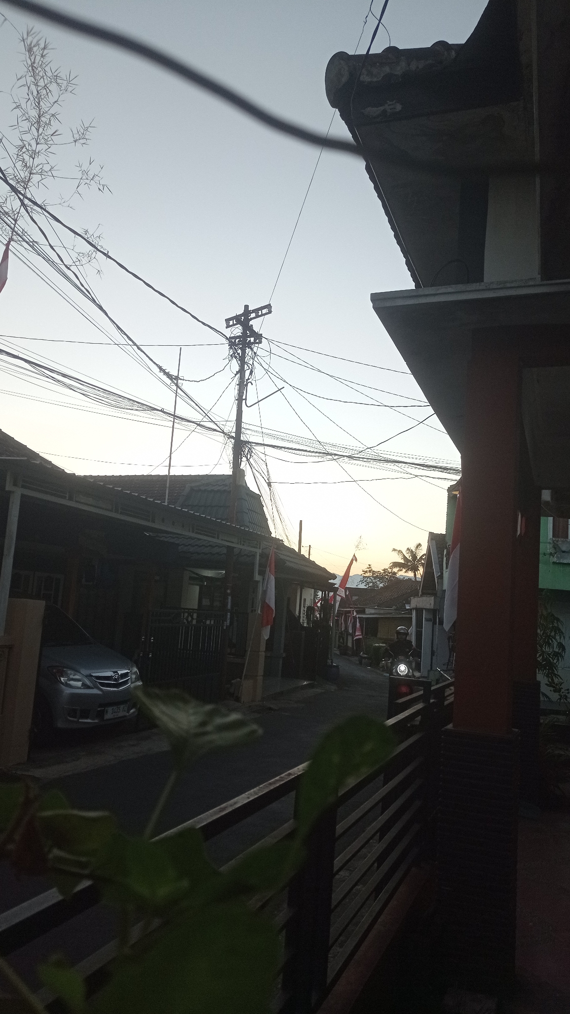 The image depicts a street scene in Mulyoagung, Indonesia, at dusk. The sky is clear and a pale yellow, indicating either sunrise or sunset. A dense network of electrical wires crisscrosses the sky, connected to a utility pole. Several houses with tiled roofs line the street, some featuring covered carports. Indonesian flags, red and white, are visible in front of some residences, suggesting a national holiday or celebration. In the foreground, a metallic gray car is parked under a carport, and a blurry green plant obstructs the lower left portion of the frame. To the right, a portion of a building with an orange pillar and a railing is visible. Further down the street, a person on a motorcycle with its headlight on is riding away from the camera. The overall impression is a quiet residential street in an Indonesian town during twilight.