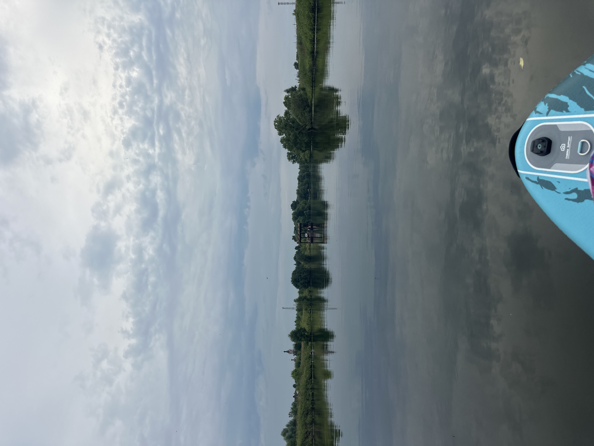 The image depicts a serene lake or wide river in Gorki Leninskiye, Russia, under a cloudy sky. The water is calm and reflects the overcast sky and the lush green trees lining the shore. On the left side of the shore, a small church with a prominent dome is visible, suggesting a rural or semi-urban setting. Further in the distance, a tall, slender tower can be seen. On the right side of the water, there is a floating wooden structure or raft with people on it, suggesting a recreational area. The foreground of the image shows the nose of a paddleboard with a camera mounted on it, indicating that the photo was taken from the perspective of someone engaged in water sports. The overall atmosphere is tranquil and peaceful, typical of a calm day on a body of water.