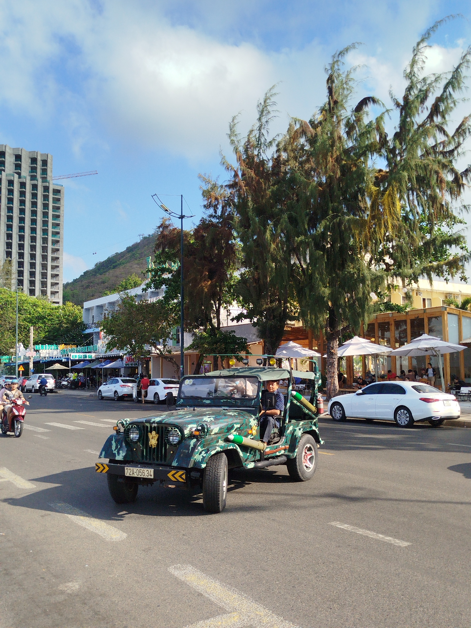 The image depicts a street scene in what appears to be a coastal area of Vietnam. A camouflage-painted vintage Jeep is prominent in the foreground, displaying a license plate with the code "72A-056.34." The driver, an adult male, is visible behind the wheel, wearing a dark-colored short-sleeved shirt. The Jeep is equipped with decorative green rocket-shaped objects on its sides.
In the background, a street with various vehicles including cars and motorbikes is visible. On the right side of the frame, there's a building with outdoor seating under white umbrellas. A tall, modern building under construction is seen in the distance on the left side of the image. The sky is bright blue with scattered clouds.