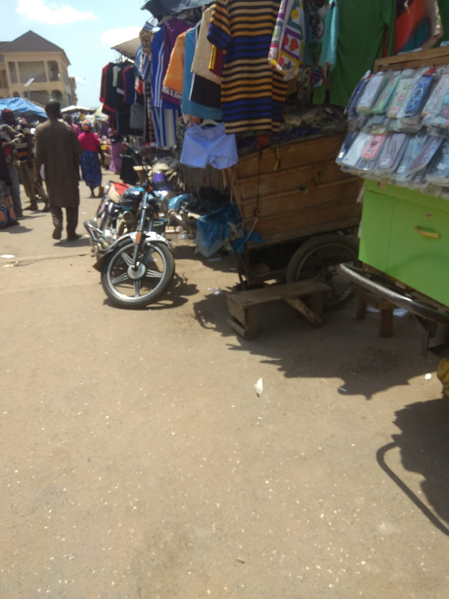 This is a street scene in Jalingo, Nigeria, likely a market or a bustling roadside area. In the foreground, a motorcycle is parked on a paved surface. Behind the motorcycle, a wooden cart is visible, with some blue fabric draped over it. To the right, a green stall or kiosk is present, displaying an assortment of items in clear plastic packaging, possibly phone cases or small accessories. Further in the background, several people are walking, and a building with multiple levels can be seen. Clothing items, including striped shirts and blue shorts, are displayed on racks or hangers, indicating a commercial setting. The overall environment appears to be outdoors, with natural daylight.