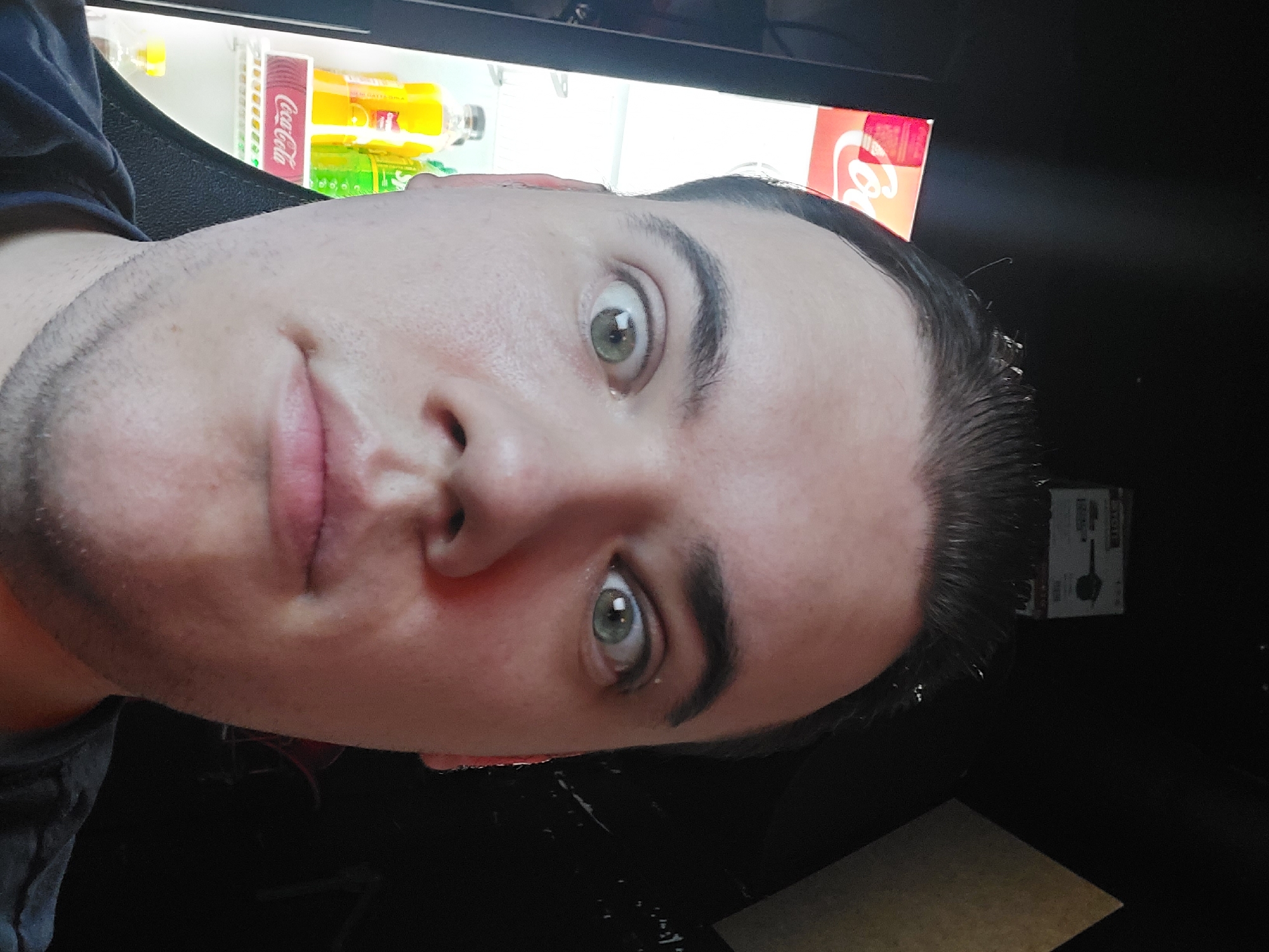 A close-up, rotated selfie captures a young person, likely male, with fair skin, dark hair, and striking green eyes. The individual looks directly into the camera with a wide-eyed, possibly surprised or intensely curious expression. Light stubble is visible on his chin and jawline, and he wears a dark-colored top.
The background is sharply divided between a brightly lit section and a very dark area. The lit section reveals the interior of a commercial refrigerator or display case, stocked with bottled beverages. Prominently visible is the red and white "Coca-Cola" logo on cans or bottles, alongside yellow and green bottled drinks. The dark background offers minimal detail, suggesting a dimly lit indoor environment around the illuminated cooler.
The scene appears to be set indoors, possibly in a store, cafe, or similar establishment in Tashkent, Uzbekistan. The overall lighting suggests it might be evening or a space with controlled, interior illumination. The activity is a self-portrait, capturing a direct and engaging moment.