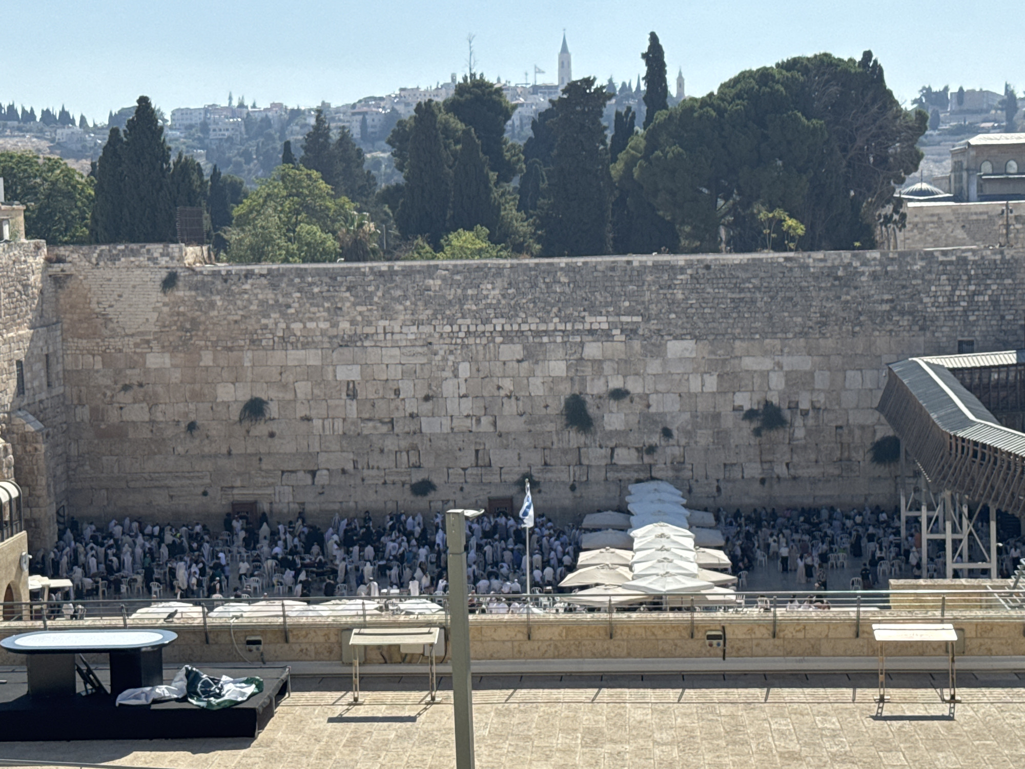 The image captures a scene at the Western Wall in Jerusalem, Israel. A large stone wall, characteristic of the ancient structure, dominates the center of the frame. In front of the wall, a multitude of people are gathered, many seated on white chairs, while others stand. They are facing the wall, suggesting a religious or prayerful gathering. Various white umbrellas are set up, providing shade for some of the attendees.
A flagpole with a blue and white flag is visible in front of the wall. To the left and right of the main gathering area, there are covered walkways or structures, one of which is a wooden walkway extending upwards. In the foreground, a low stage-like structure is set up with a dark platform and a white, oval-shaped table. A green and white cloth or flag is draped over the platform. The surrounding environment includes trees and buildings on a hillside in the background, indicative of the city's landscape. The sky is clear and blue, suggesting a sunny day.
