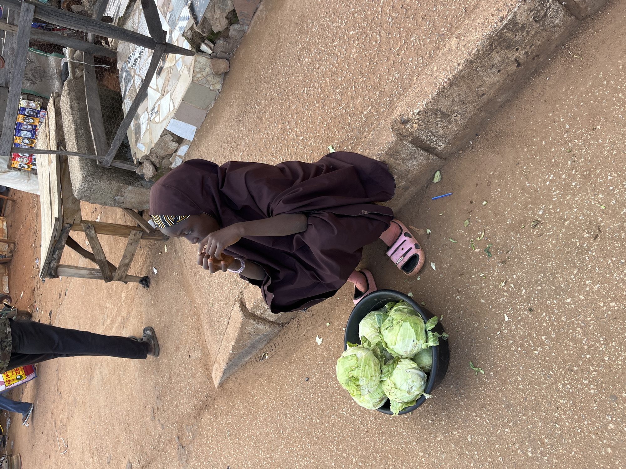 In Bukuru, Nigeria, a young person is seated on the ground by the roadside, wearing a dark brown hijab and robe. They are holding what appears to be a piece of food to their mouth. In front of them, a black bucket is filled with several heads of fresh green cabbage. The ground is a mix of reddish-brown dirt and small pebbles, with a concrete curb bordering the street. In the background, a wooden market stall is visible, with various items displayed on shelves. The scene suggests a bustling market environment.