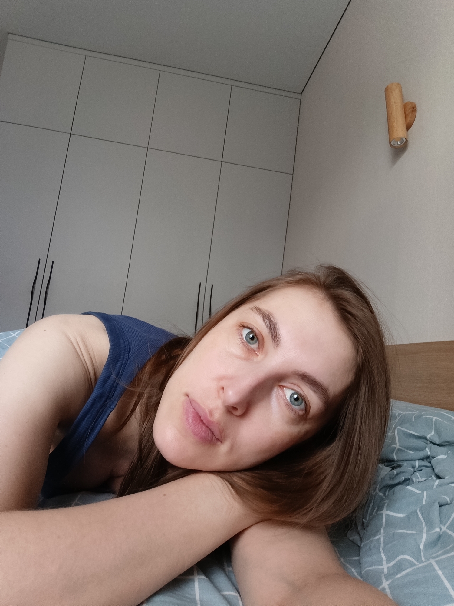 A young woman is pictured lying down in what appears to be a modern bedroom in Chaikovsky, Russia. She is positioned on her side, resting her head on her folded arms which are propped on a light blue duvet patterned with a white grid. She looks directly at the camera with a calm and relaxed expression.
The woman has light skin, blue-grey eyes, and long brown hair. She is wearing a simple blue tank top. The background features a minimalist interior design. Behind her is a large, light grey modular wardrobe or cabinet system with thin black lines delineating rectangular panels, and subtle black vertical handles on some sections. To her right, a plain beige wall hosts a modern, cylindrical wooden wall sconce. The lighting is soft and diffused, suggesting daytime. There is no visible text in the image.