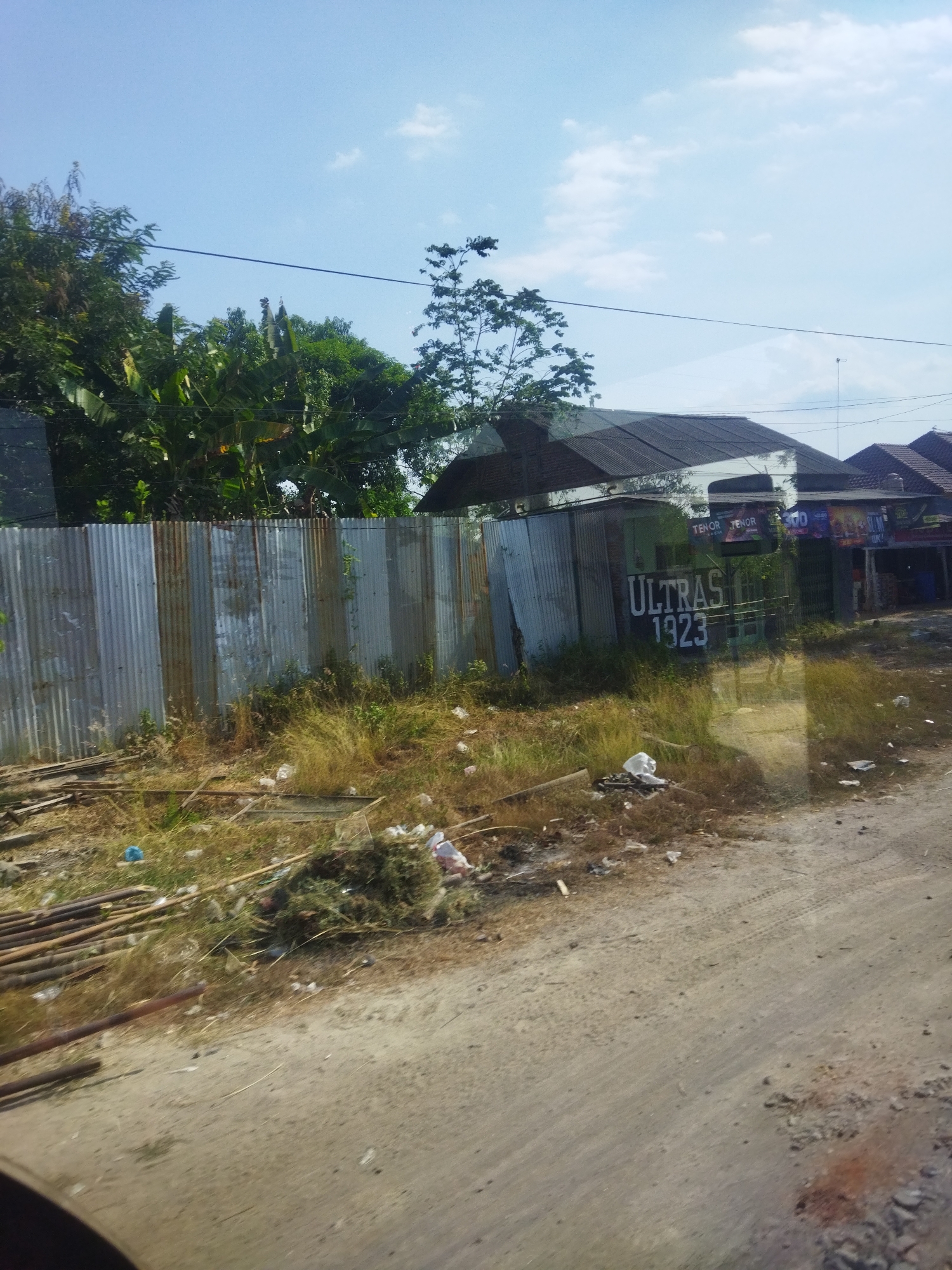 The image depicts a roadside scene in Nguter, Indonesia. A corrugated metal fence runs along the left side of the frame, partially obscuring a lush green area with trees and vegetation. In front of the fence, dry grass and scattered debris, including plastic bags and wooden poles, are visible on the dusty ground. Further in the background, several buildings with dark-tiled roofs are present. One building has "ULTRAS 1923" painted in large white letters on its side. The sky is clear and blue with a few scattered white clouds. The overall impression is one of a somewhat unkempt, semi-rural or suburban environment under a bright sky.