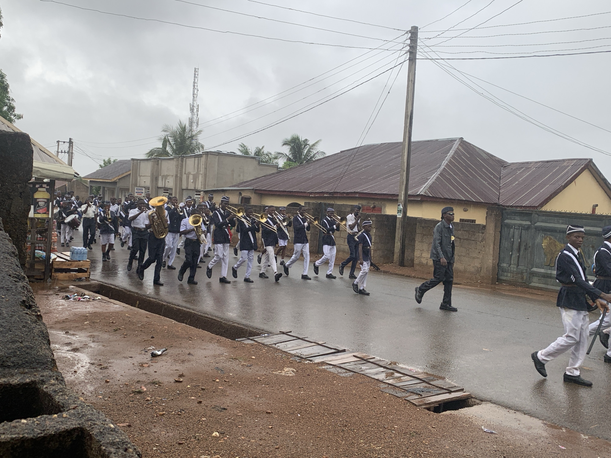 A street parade takes place in Akwanga, Nigeria. A marching band of several dozen people in matching uniforms walks along a wet asphalt street, bordered by open storm drains covered with wooden planks. Most band members wear navy-blue jackets, white pants, and matching hats. Most are carrying and playing brass instruments. Some are marching with drum kits. Buildings and utility poles line the street. The sky is overcast.