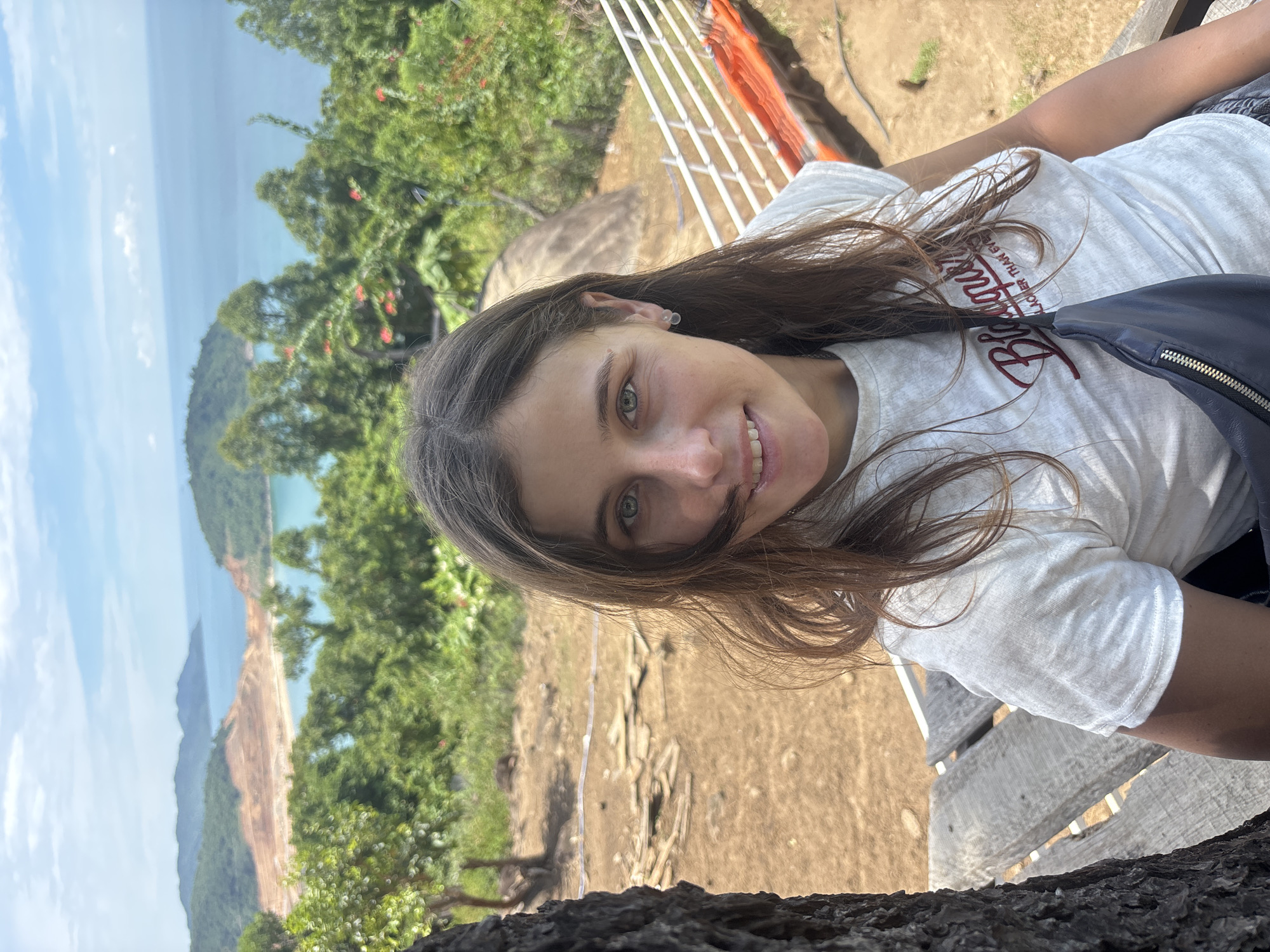 The media file is a portrait of a young woman outdoors. She is positioned in the foreground, looking towards the camera with a slight smile and her head tilted. Her long, brown hair is loose and falls around her face and shoulders. She is wearing a white t-shirt with a graphic design on the front, which appears to be a stylized word in dark red lettering. A dark blue fanny pack is visible around her waist.
The background depicts a scenic view of a coastline. Lush green trees and foliage dominate the mid-ground, with a distinct strip of tan, rocky terrain leading down to a body of water, likely the ocean. The water is a pale blue, and distant hills or mountains are visible, shrouded in a light haze. The sky above is a bright blue with scattered white clouds. The overall impression is of a sunny day in a tropical or subtropical location.
The composition suggests the photograph was taken from a slightly elevated position, possibly on a viewing platform or a high point overlooking the coast. The presence of wooden planks and a white railing in the upper right corner further supports this. The woman's relaxed posture and pleasant expression convey a sense of enjoyment and perhaps a vacation. The image appears to be a selfie or a photo taken by someone accompanying her. The visible text on her shirt is not fully legible due to the resolution and angle. The location context suggests this scene is in Đà Nẵng, Vietnam.