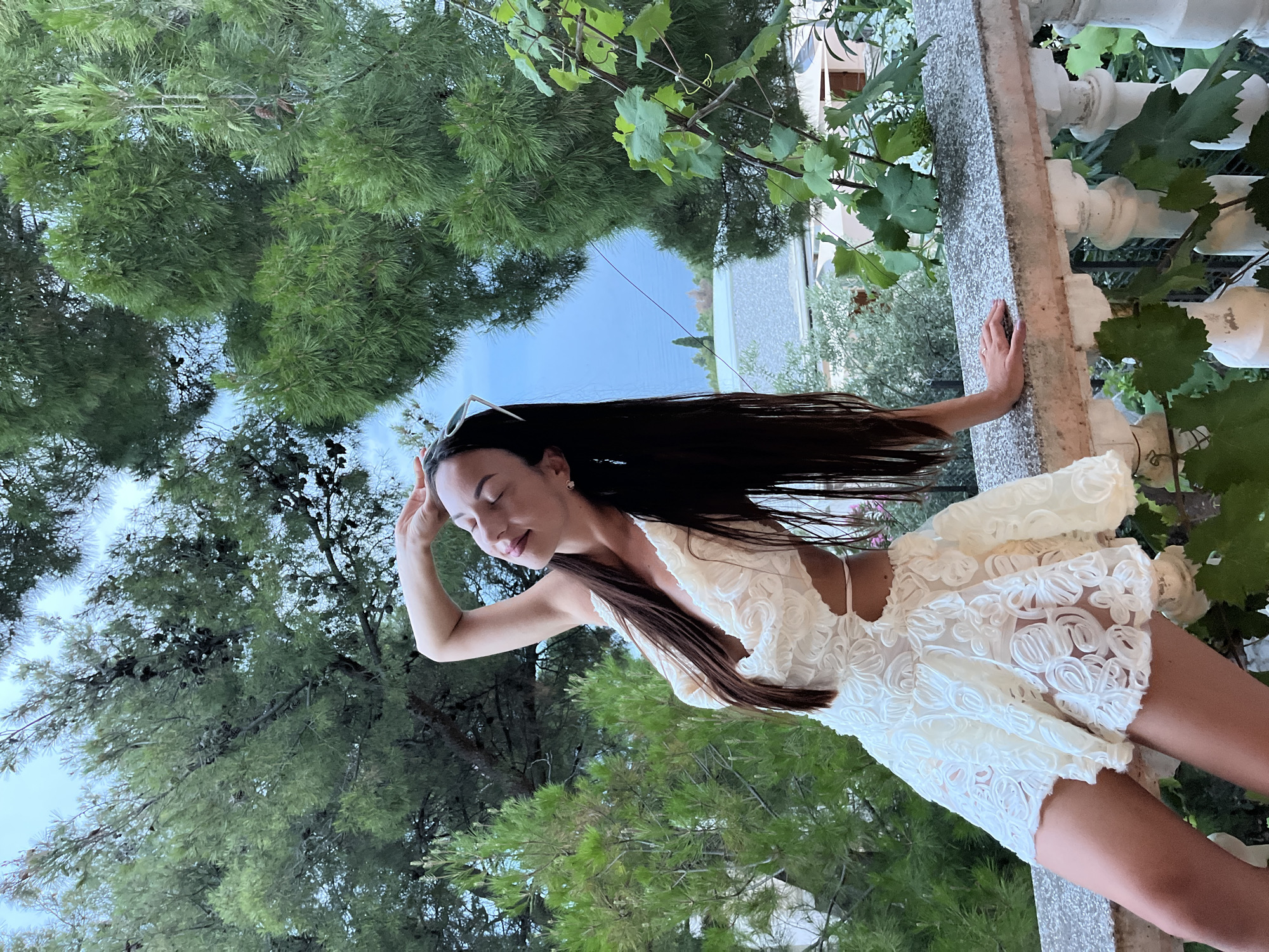 A woman with long dark hair, wearing a white lace dress, leans on a stone balustrade overlooking a body of water. She has her right hand on her forehead, her eyes closed, and her head tilted back slightly, suggesting relaxation or enjoyment of the sun. The dress features cutouts at the sides and a plunging neckline. Behind her, lush green trees and foliage frame the scene, with buildings and a coastline visible in the distance. The setting appears to be a balcony or terrace in a coastal town, potentially Šušanj, Montenegro, given the Mediterranean vegetation and architecture.