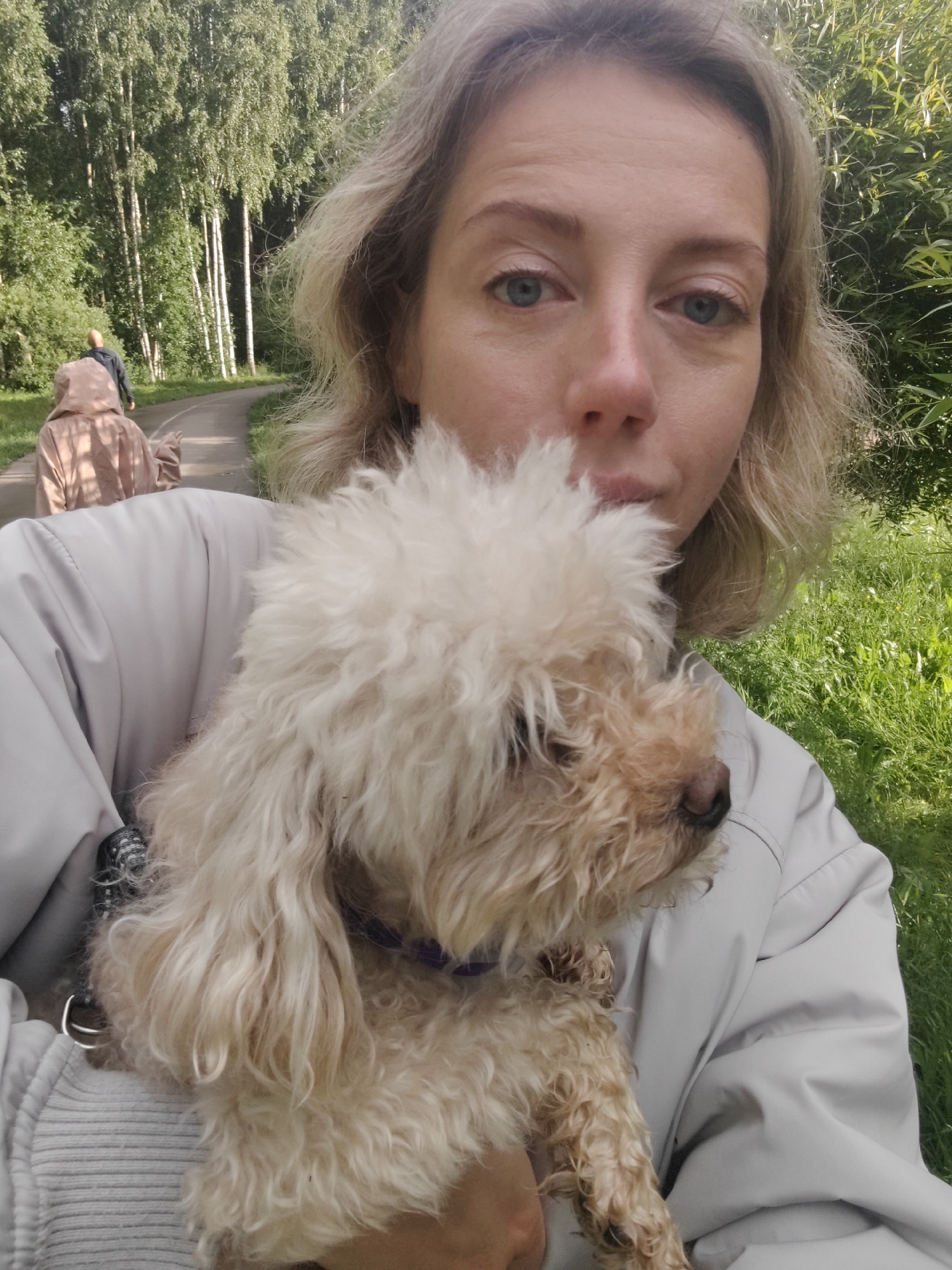 A blonde woman is holding a small, light-colored poodle in her arms. She is wearing a light-colored jacket. The dog has curly fur. In the background, a paved path curves through a park lined with trees. Two figures are walking on the path in the distance. The ground is covered with grass. The scene is outdoors during the daytime.