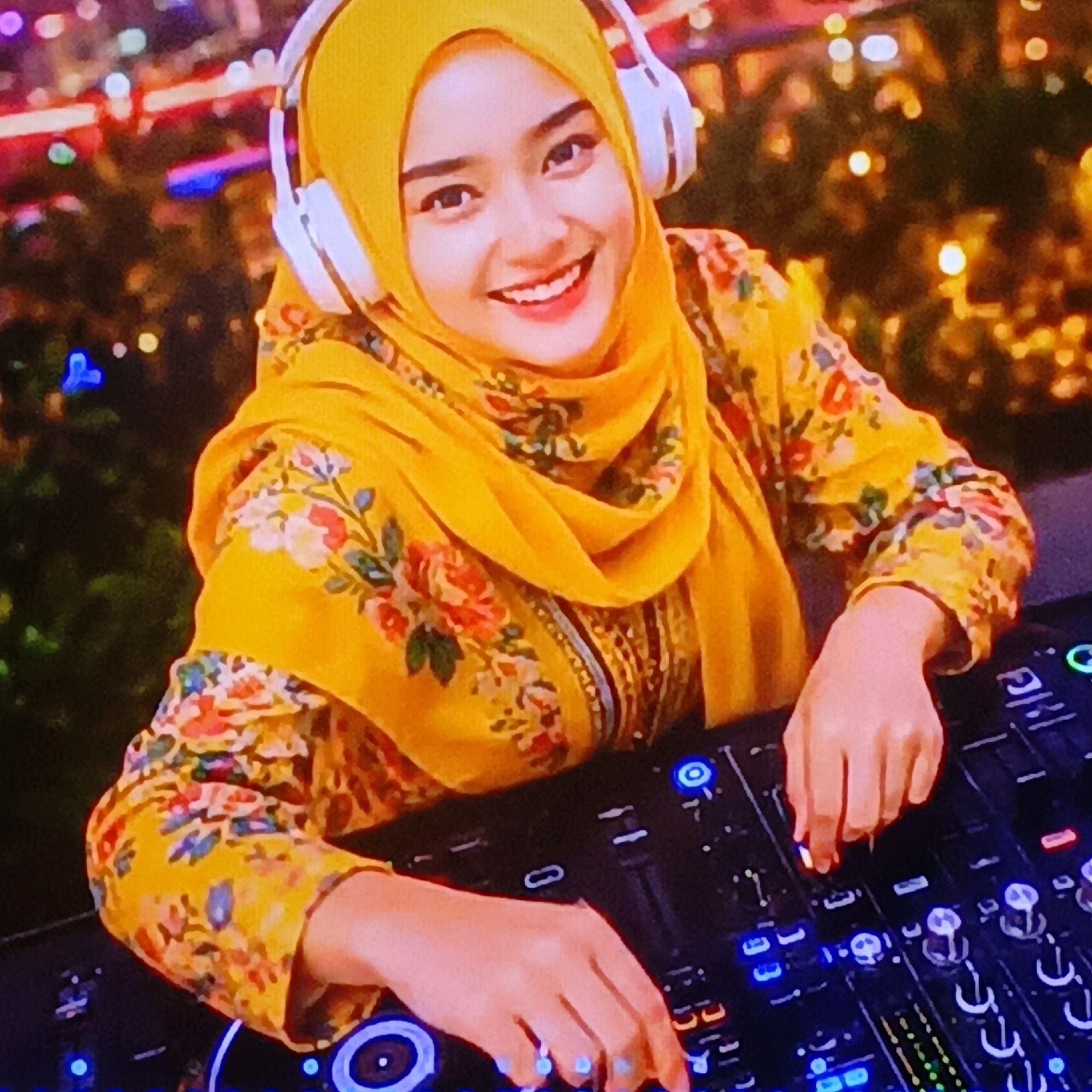The image shows a young woman wearing a yellow hijab and a yellow floral-patterned top. She is also wearing white headphones and is in front of a DJ console. Her hands are on the DJ console, with one hand operating a slider. The background is blurred and appears to be a city at night, with bokeh lights. The scene is captured from a medium shot, focusing on the woman and the DJ equipment. The woman is smiling and looking towards the camera. The environment suggests an indoor or outdoor event at night with a vibrant atmosphere, possibly a club or an outdoor festival in Salatiga, Indonesia.