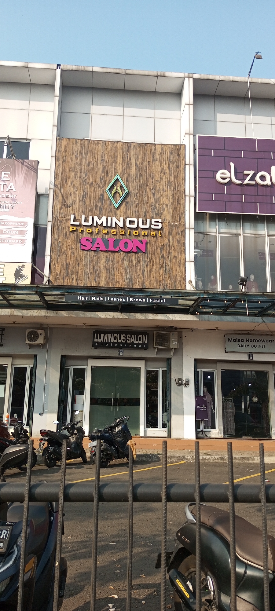 The image shows a street view of the "LUMINOUS PROFESSIONAL SALON" located in Lambangjaya, Indonesia. The salon's signage is prominently displayed on a wooden-paneled facade above the entrance. Below the main sign, a smaller strip sign lists services offered: "Hair | Nails | Lashes | Brows | Facial".
The salon itself has a grey exterior with glass doors and windows. Two air conditioning units are mounted on the wall above the entrances. Parked in front of the salon are several motorcycles, including a dark blue one with a tan piece of fabric draped over its rear. To the right of the salon is another storefront labeled "Maisa Homewear DAILY OUTFIT". Further to the right, a purple sign for "elzatta 2nd FLOOR" is visible.
The scene is illuminated by daylight, and the sky is clear blue. The foreground of the image is partially obscured by a black metal fence. The overall impression is of a commercial street in a suburban or urban area.