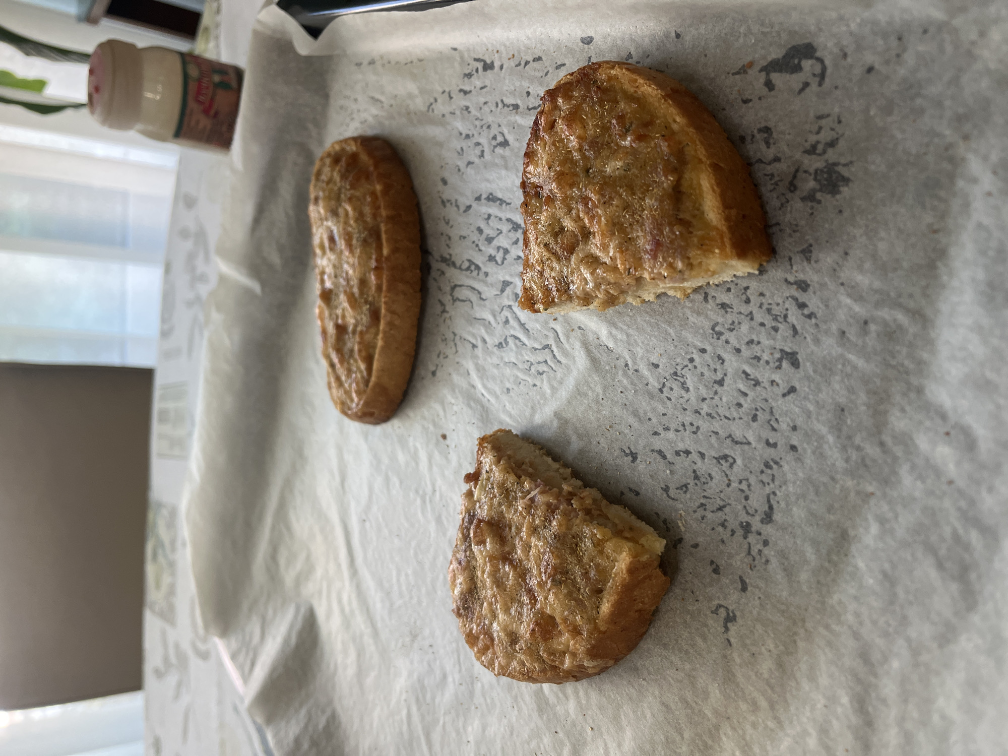The image captures three pieces of savory toasted bread, possibly garlic bread or a similar appetizer, resting on white parchment paper laid on a baking sheet. The bread is golden brown, and each piece is topped with a melted, slightly browned mixture that appears to include herbs and possibly cheese. One piece is a full, elongated slice, another is a half-slice, and the third is a smaller, irregular triangular piece. The parchment paper beneath the bread shows various dark, speckled marks, suggesting it may have been used for baking.
In the upper left corner of the frame, a white plastic shaker bottle with a red cap is partially visible. The label on the bottle is clear enough to show the brand name "DeSantis" and the content "Origano." The background is softly blurred but reveals a bright, naturally lit interior space with a window and light-colored curtains, indicating it is daytime.
The scene suggests a domestic setting, likely a kitchen or dining area in Sofia, Bulgaria, where food is being prepared or has just been finished. The bread appears to be freshly made and ready to be eaten, or awaiting further cooking.