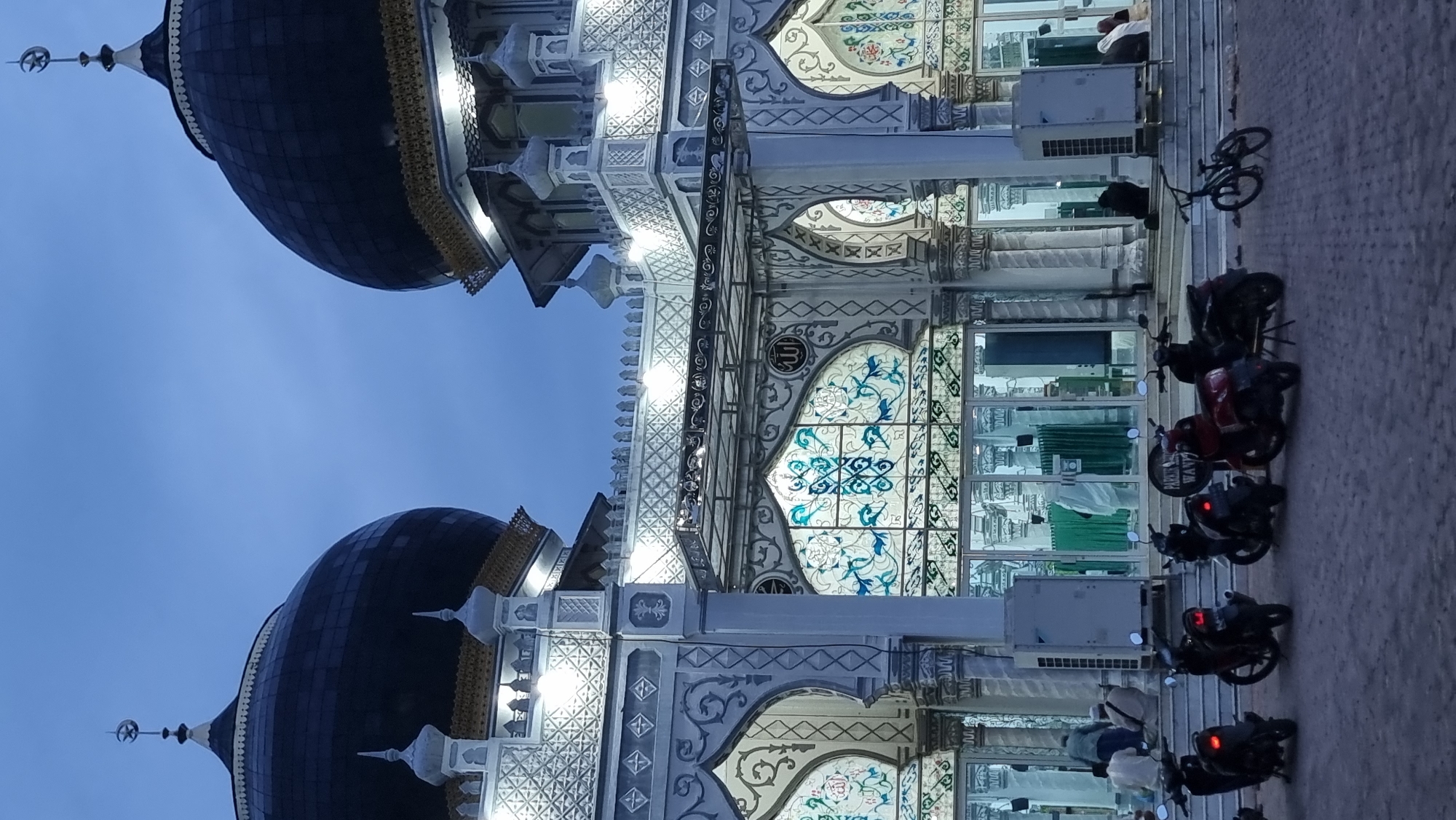 The image depicts the exterior of a mosque in Keudekruenggeukueh, Indonesia, during twilight. Two prominent dark domes, topped with crescent moons, dominate the upper portion of the frame. The mosque features intricate architectural details, including patterned facades with arched windows filled with stained glass in blue and white hues. White decorative elements and gold accents are visible on the building's exterior. Several individuals are seen near the entrance, with some seated on steps and others standing. The scene is illuminated by artificial lights that highlight the mosque's architecture. In the foreground, several motorcycles and a bicycle are parked on the paved ground in front of the mosque. The overall ambiance suggests a serene, possibly evening, scene at a place of worship.