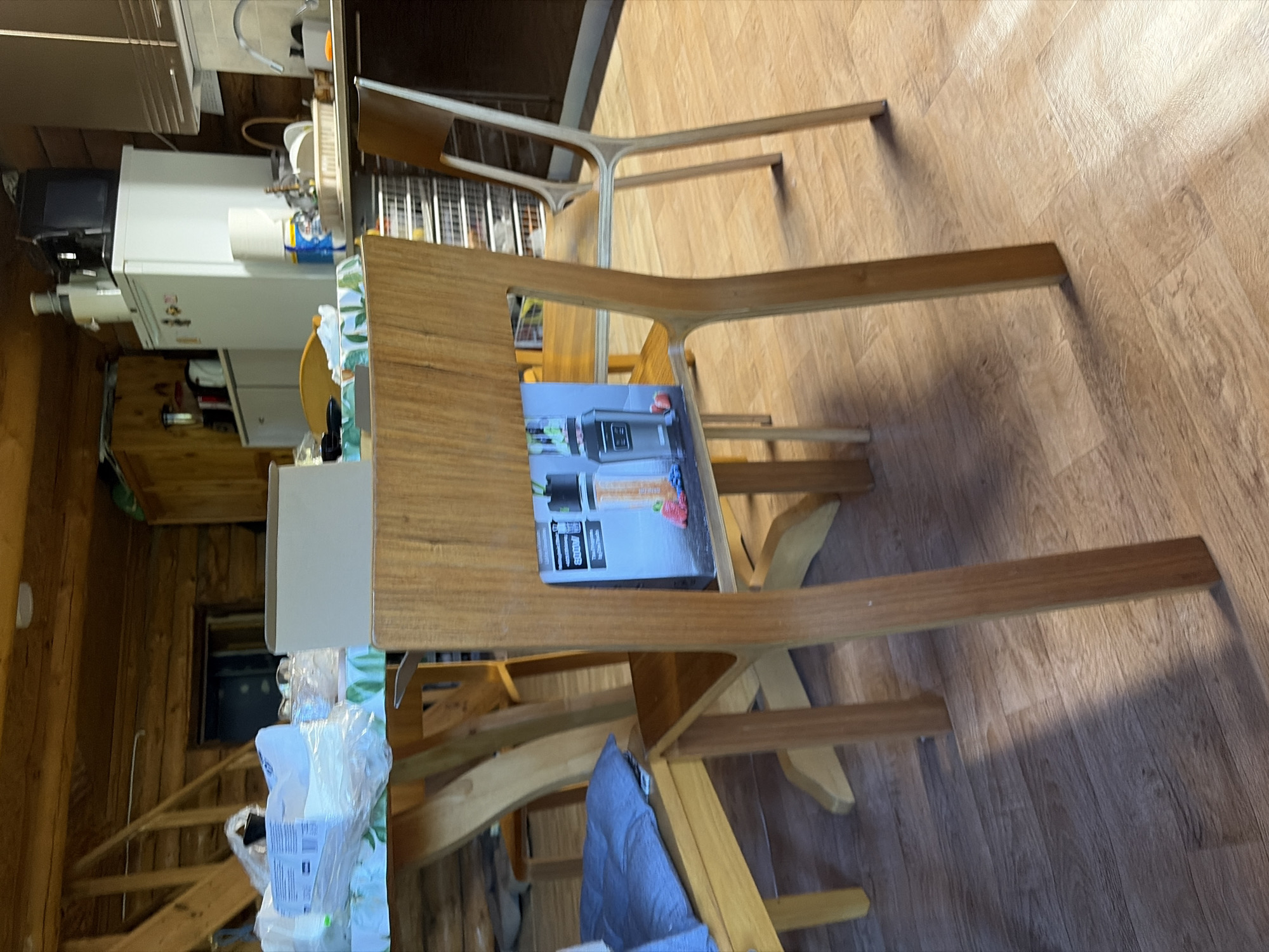 The image shows a wooden dining table with a light-colored wood grain, placed on a light wood-patterned floor. The table has a unique, curved design, with its legs extending upwards and forming part of the tabletop structure. On the table, there is a product box for a Ninja Foodi blender, featuring an image of the appliance and its accessories. To the left of the table, part of a white kitchen appliance, possibly a refrigerator or a large food processor, is visible. Beyond that, a wooden staircase leads upwards, and the walls are made of rough-hewn wood logs, suggesting a rustic or cabin-like setting. A blue cloth is draped over a chair or stool under the table. The lighting suggests it is daytime, with shadows indicating light sources from the right and above. The scene appears to be indoors, likely in a kitchen or dining area of a residential property in Stīveri, Latvia, given the provided location context. There are no people visible in the image, and the overall mood is calm and undisturbed. Visible text includes "Ninja Foodi" on the product box.