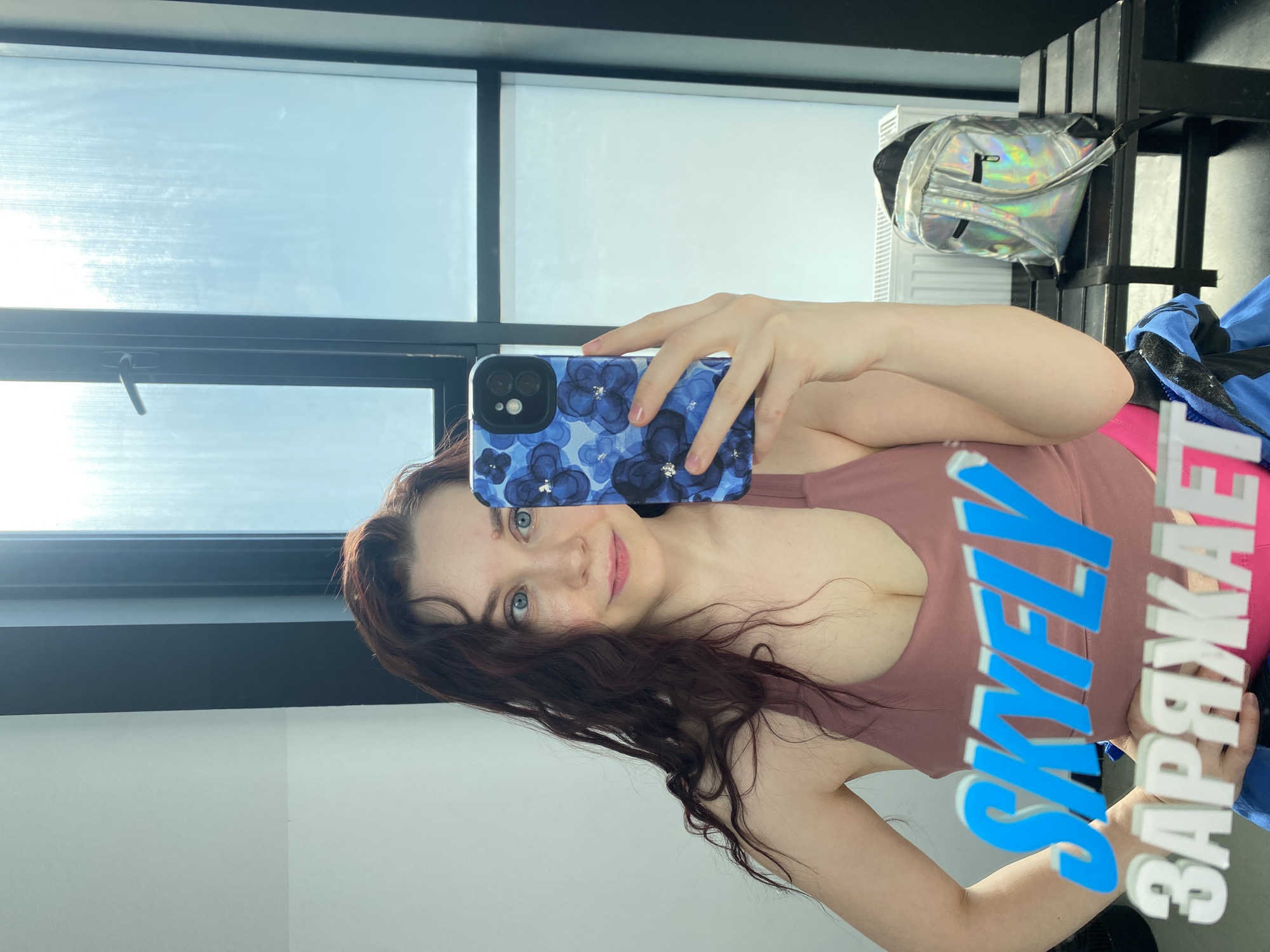 A woman is taking a selfie in front of a window. She is holding a phone with a blue floral case. She is wearing a mauve tank top. Her hair is dark and wavy. There is a silver backpack on a shelf behind her. A sticker with the word "SKYFLY" in English and "ZARJAE" in Russian is superimposed on the image. The location is possibly in Izhevsk, Russia, given the Russian text.