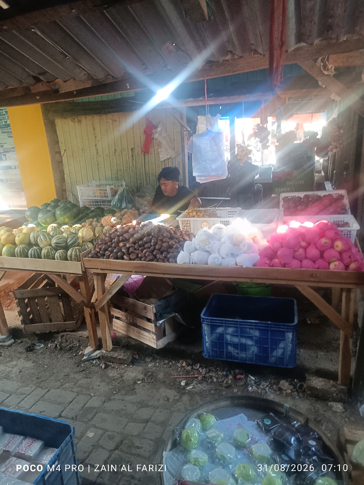 The image depicts an outdoor market stall in Arosbaya, Indonesia, showcasing a variety of fruits. In the foreground, a wooden table is laden with several types of fruit. To the left, there are rows of round, green and yellow fruits, likely melons, and behind them, larger dark green watermelons. To the right of the melons, a pile of dark brown, oval-shaped fruits, possibly salak, are displayed. Further to the right, on another section of the table, are white-wrapped fruits and a pile of pink-wrapped apples. In the background, a man in a dark shirt is visible behind the fruit stall, appearing to interact with the produce. Behind him, various items, including plastic bags and white crates, are present. The market stall has a rustic roof made of corrugated metal supported by wooden beams, and the walls are constructed from vertical wooden planks. The ground is covered with dirt and some cobblestone paving. A blue plastic crate is placed on the ground to the right, and another wooden crate is visible beneath the table on the left. In the lower portion of the image, a circular metal basin holds more green fruits, some of which are individually wrapped. The timestamp "31/08/2026 07:10" and text "POCO M4 PRO | ZAIN AL FARIZI" are overlaid on the bottom of the image.