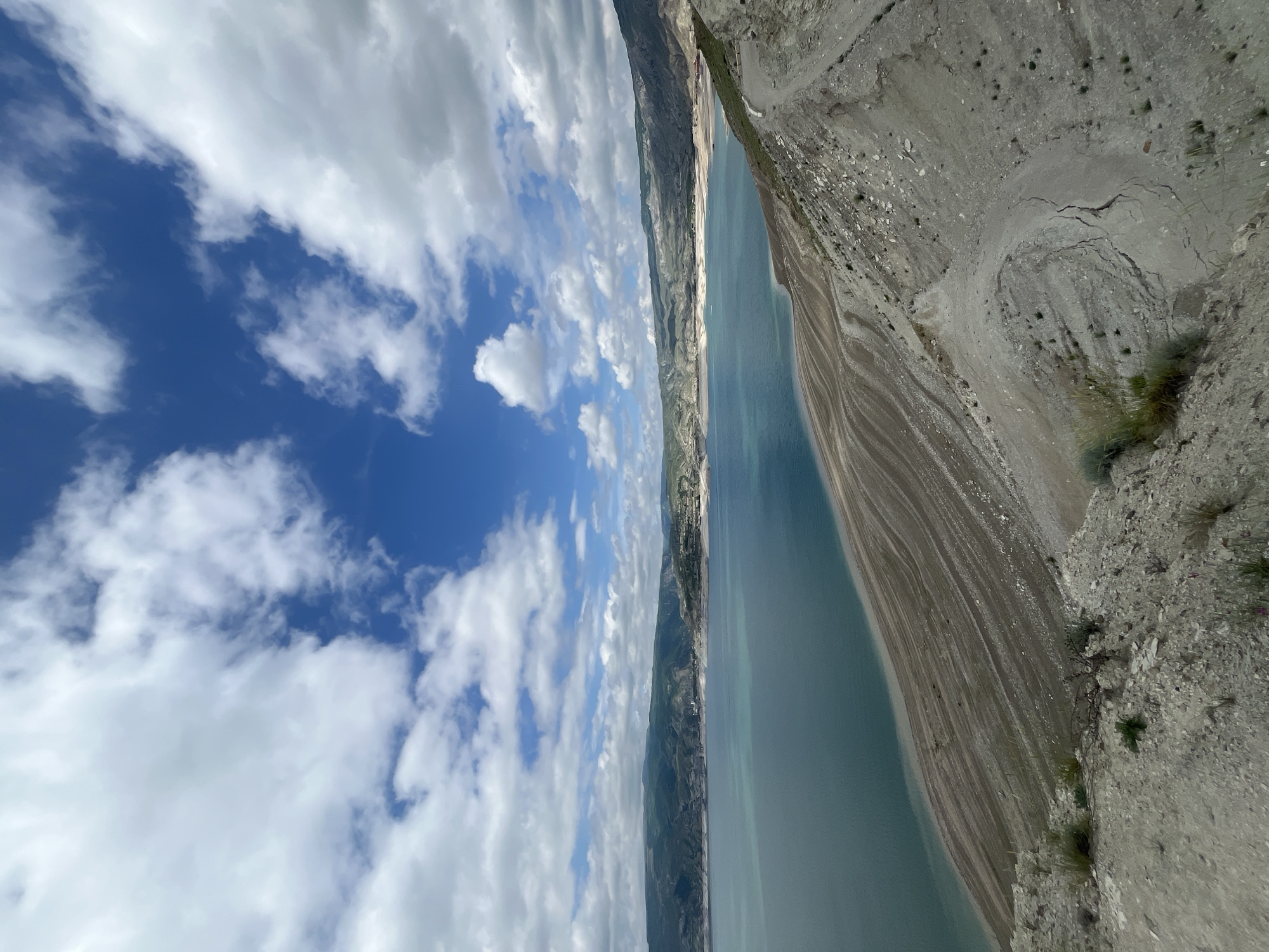 The image presents an aerial view of a landscape. On the right side, there is a rocky, mountainous terrain with sparse vegetation. A body of water, resembling a lake or reservoir, extends lengthwise along the image, and the sky above is filled with blue patches and fluffy white clouds. The location is Chirkey, Russia.