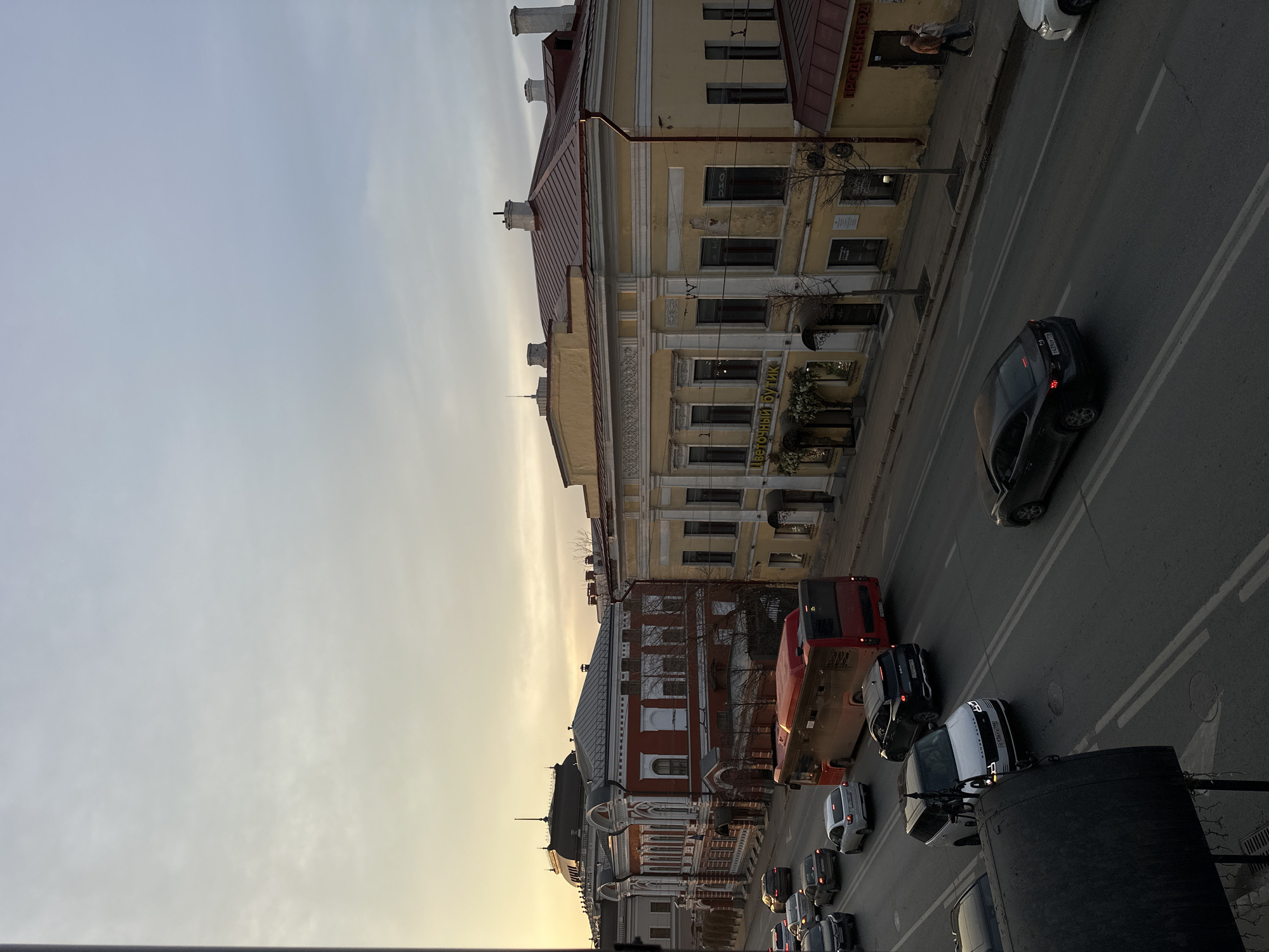 The image captures a street scene in Kazan, Russia. Buildings with distinct architectural styles line the street. A yellow building with a red roof is present, along with other buildings of varying heights and designs. Traffic includes cars and a red bus. The sky is a mix of gray and yellow, suggesting either early morning or late afternoon. The scene is viewed from an elevated perspective.
