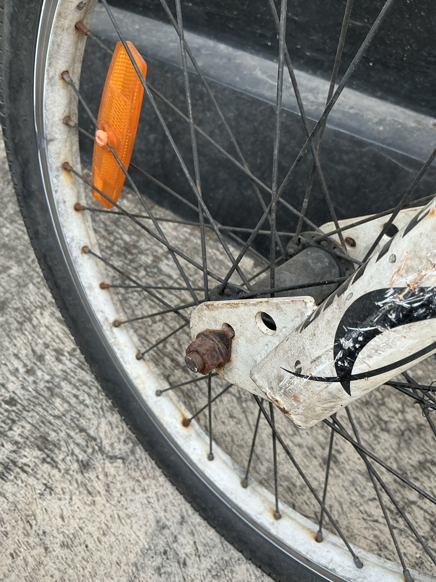 A close-up, eye-level shot focuses on the front wheel of a bicycle, resting on a concrete surface. The bicycle appears weathered, with visible rust on the spokes and a worn, white frame adorned with a black graphic. An orange reflector is attached to some of the spokes on the left side of the wheel. The background is dark and indistinct, suggesting it could be a wall or a shadow. This image was taken on Jalan Pahlawan, Magelang, Indonesia.