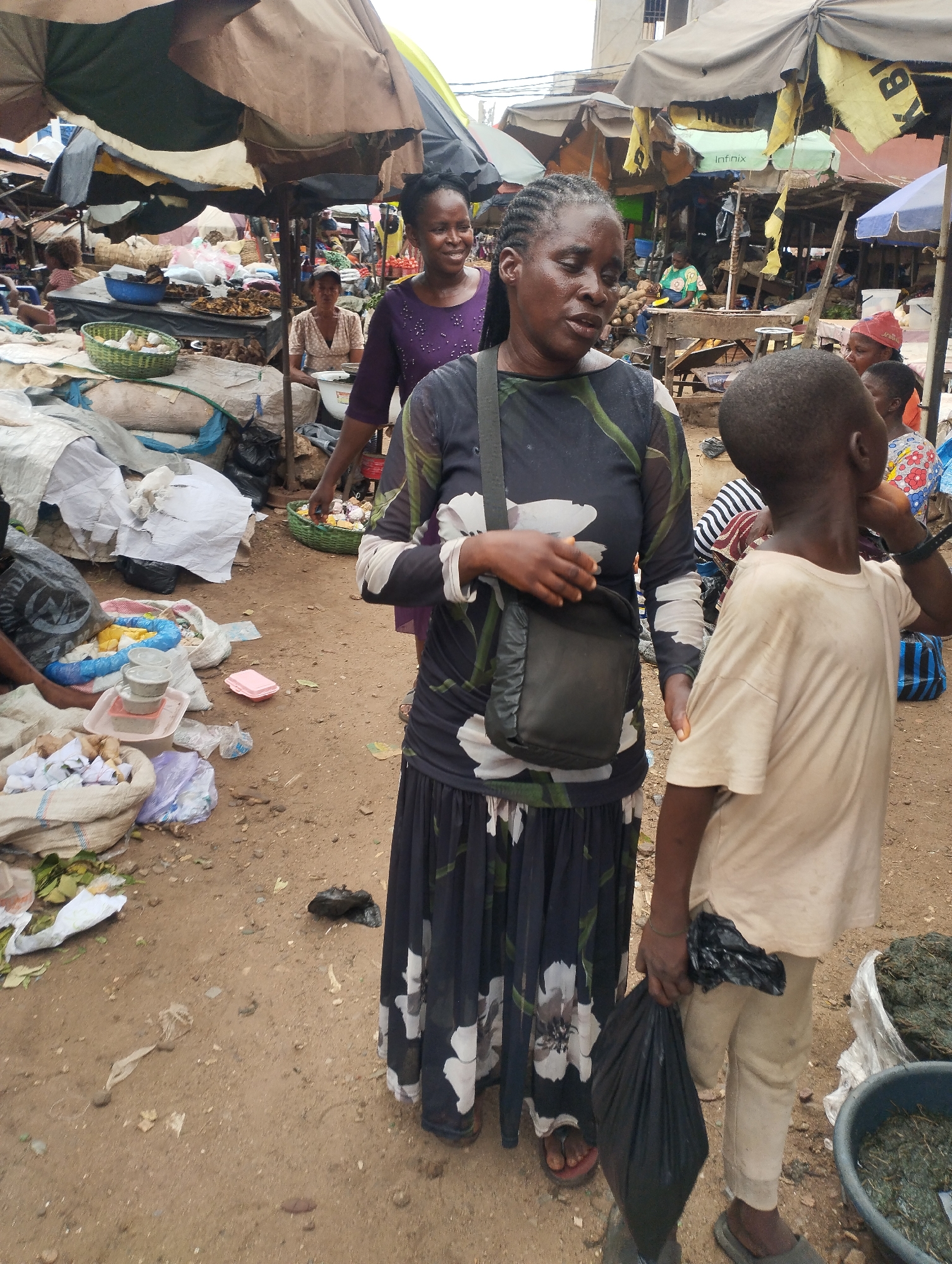 The image depicts a bustling market scene in Fegge, Nigeria. The foreground is dominated by a woman with braided hair, wearing a black and white floral dress and a dark shoulder bag, engaged in conversation. A young boy stands beside her, holding a black plastic bag. The ground is dirt-covered and littered with debris. Numerous stalls are visible under umbrellas, offering a variety of goods. Other people are scattered throughout the market, some selling items, others browsing. Bags and containers with various goods are on display on the ground.