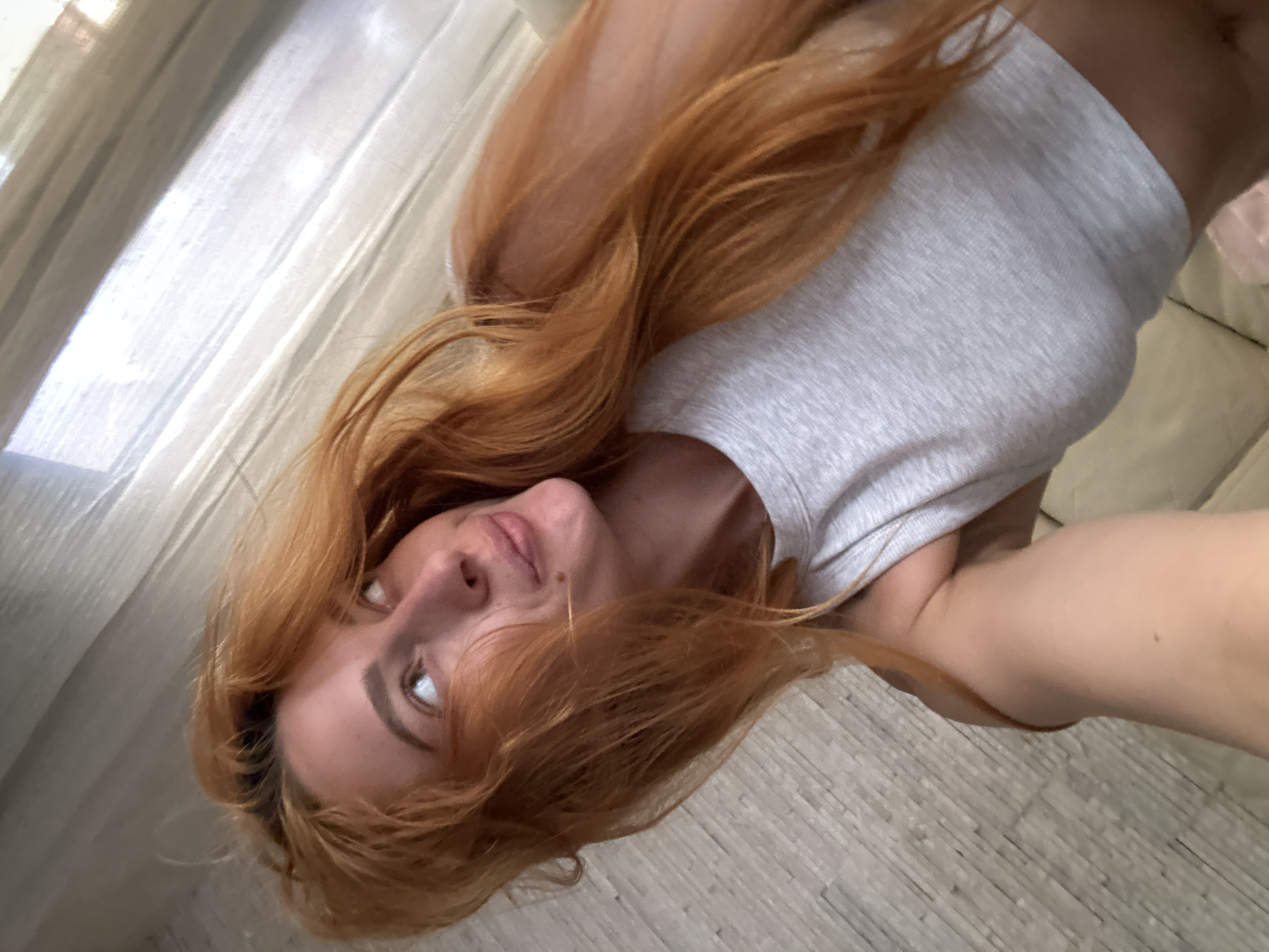 A young woman with long, auburn hair is lying down, looking up and to her left. She is wearing a light gray tank top. Her hair is spread out around her head and shoulders. The background appears to be an indoor setting, with a textured wall to her left and a sheer curtain to her right, suggesting she is near a window. The lighting is soft and natural, possibly from the window. The image captures a candid moment, with the woman appearing relaxed and contemplative. There is no identifiable location in the city of Budva, Montenegro.