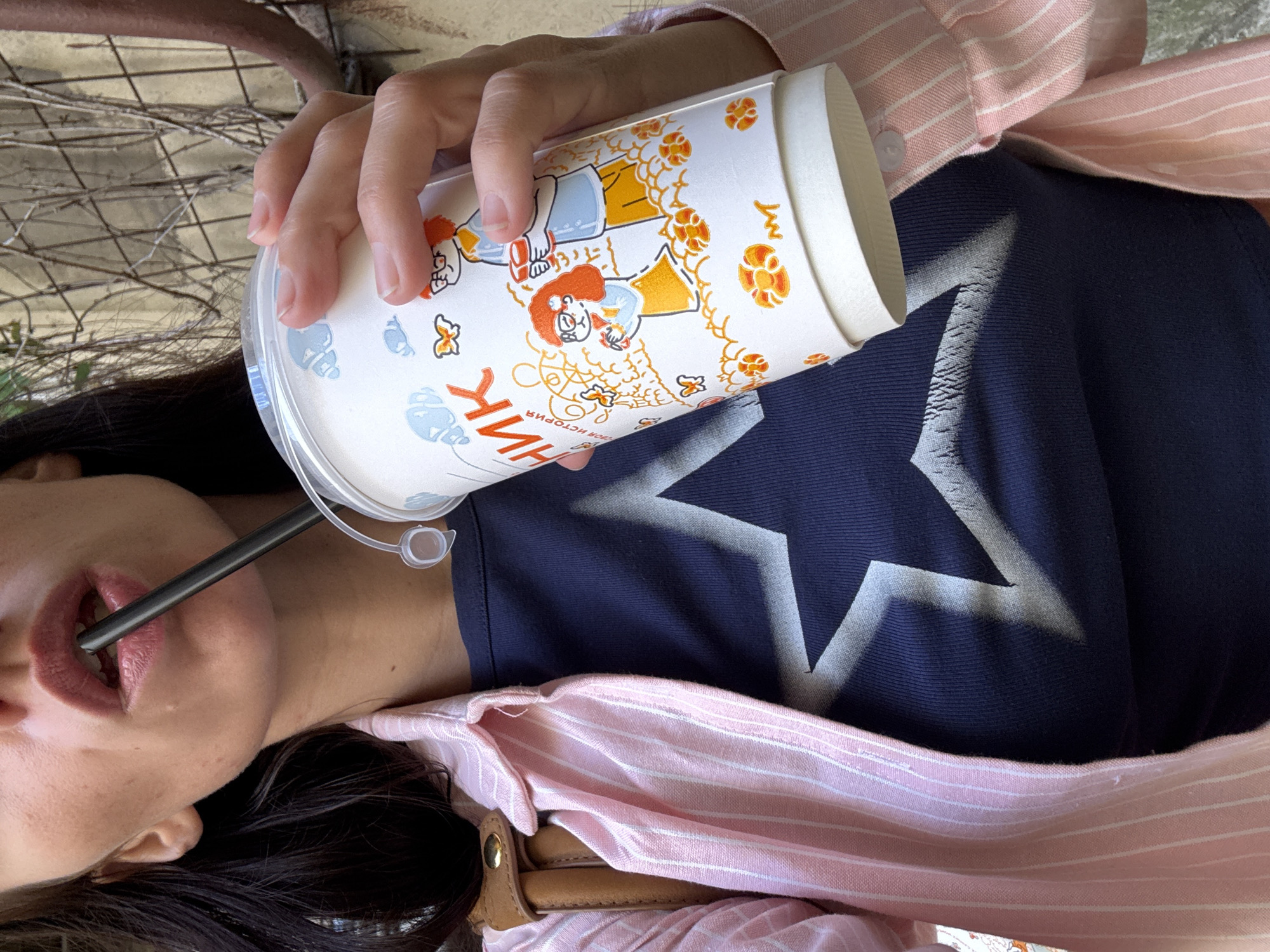 A close-up, waist-up shot shows a woman holding a disposable cup with a black straw to her lips, sipping from it. She is wearing a navy blue t-shirt with a large white star graphic and a pink and white striped button-up shirt over it. Her hair is dark brown. The background appears to be an outdoor urban setting, possibly a side street in Saint Petersburg, Russia, with a weathered, light-colored wall and some sparse, dry greenery visible on a metal fence or trellis. The lighting suggests it is daytime. The cup has cartoonish illustrations of people and butterflies.