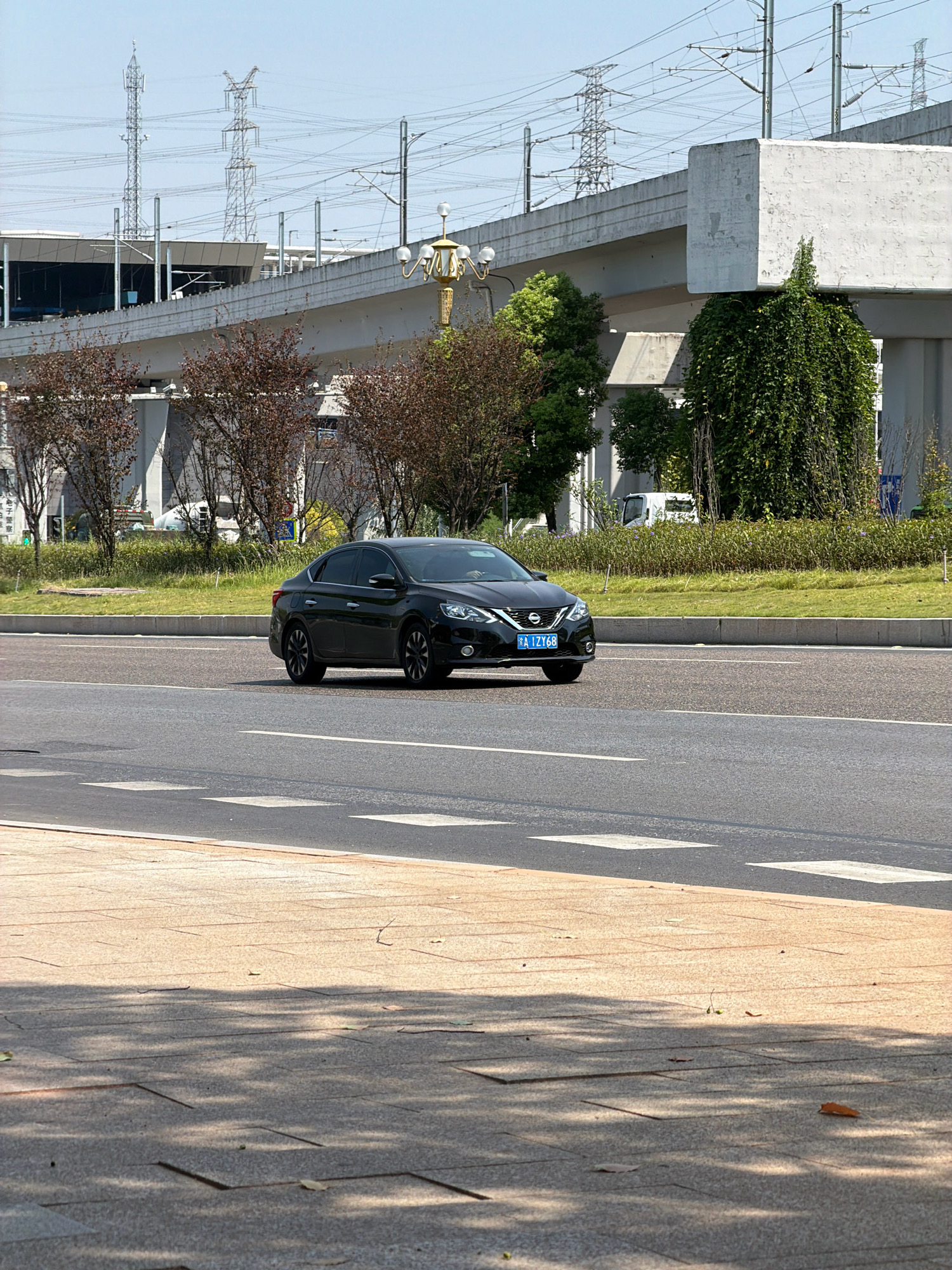 The image shows a black sedan with license plate "A IZY68" driving on a paved road in Jiangjin, China. The car is in the right lane of a multi-lane road, with a pedestrian walkway on the left side of the frame. In the background, there is an elevated structure that appears to be part of a transportation network, possibly a railway or an overpass, supported by concrete pillars. Power lines and transmission towers are visible in the distance. There are trees and landscaping along the roadside. The sky is clear and blue, indicating daytime. The scene depicts an urban or suburban environment with infrastructure for transportation and power distribution.