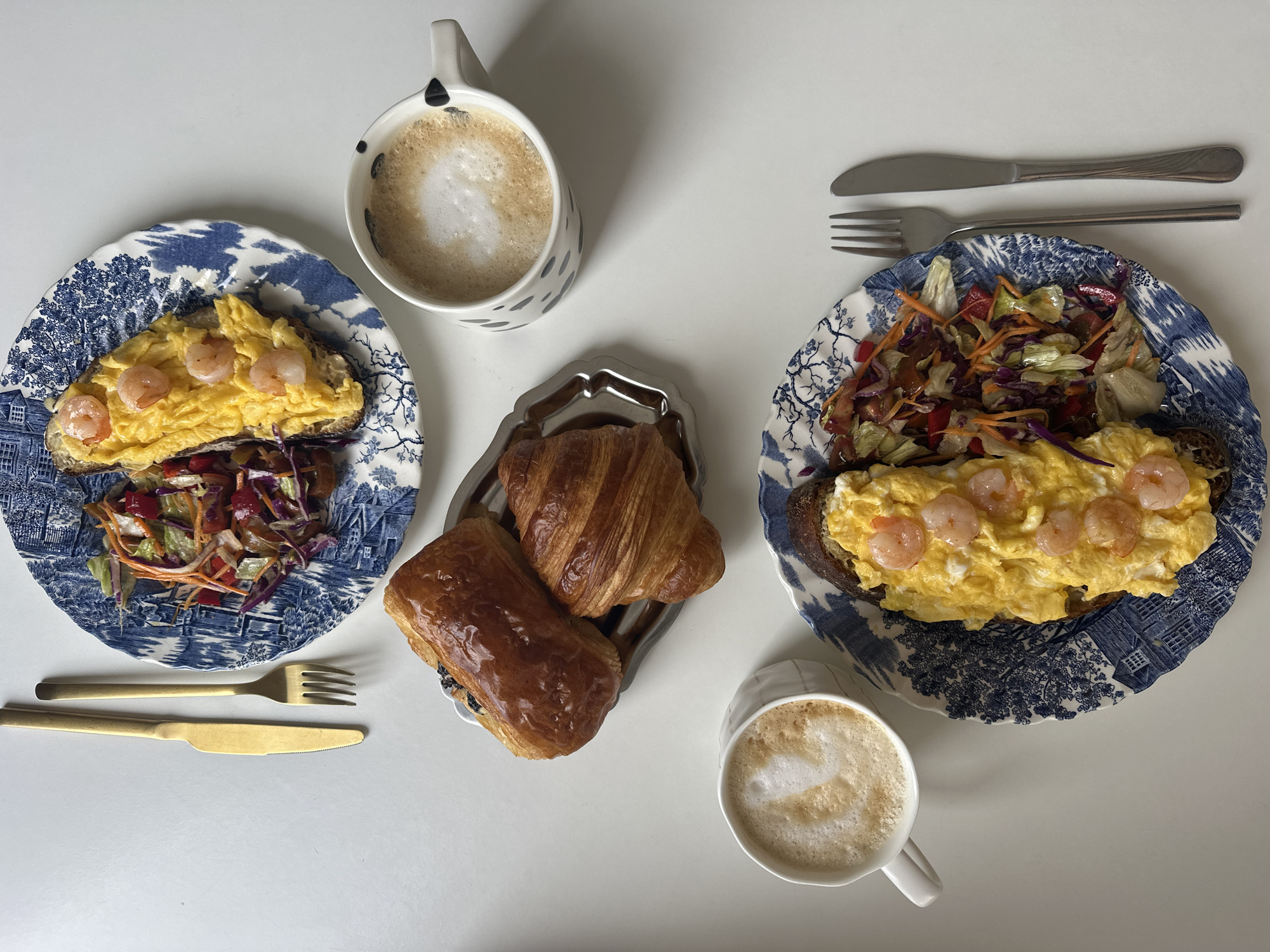 A top-down view captures a breakfast spread on a white surface. Two patterned blue and white plates are prominently displayed, each holding a slice of toast topped with scrambled eggs and shrimp, accompanied by a salad. A tray in the center contains a croissant and a pain au chocolat. Two mugs filled with coffee and foam are also present, one on either side of the tray. A gold-colored knife and fork are placed beside the plate on the left, while a silver knife and fork are positioned by the plate on the right. The scene does not suggest a specific location in Ramat Gan, Israel; it appears to be a simple breakfast setting.