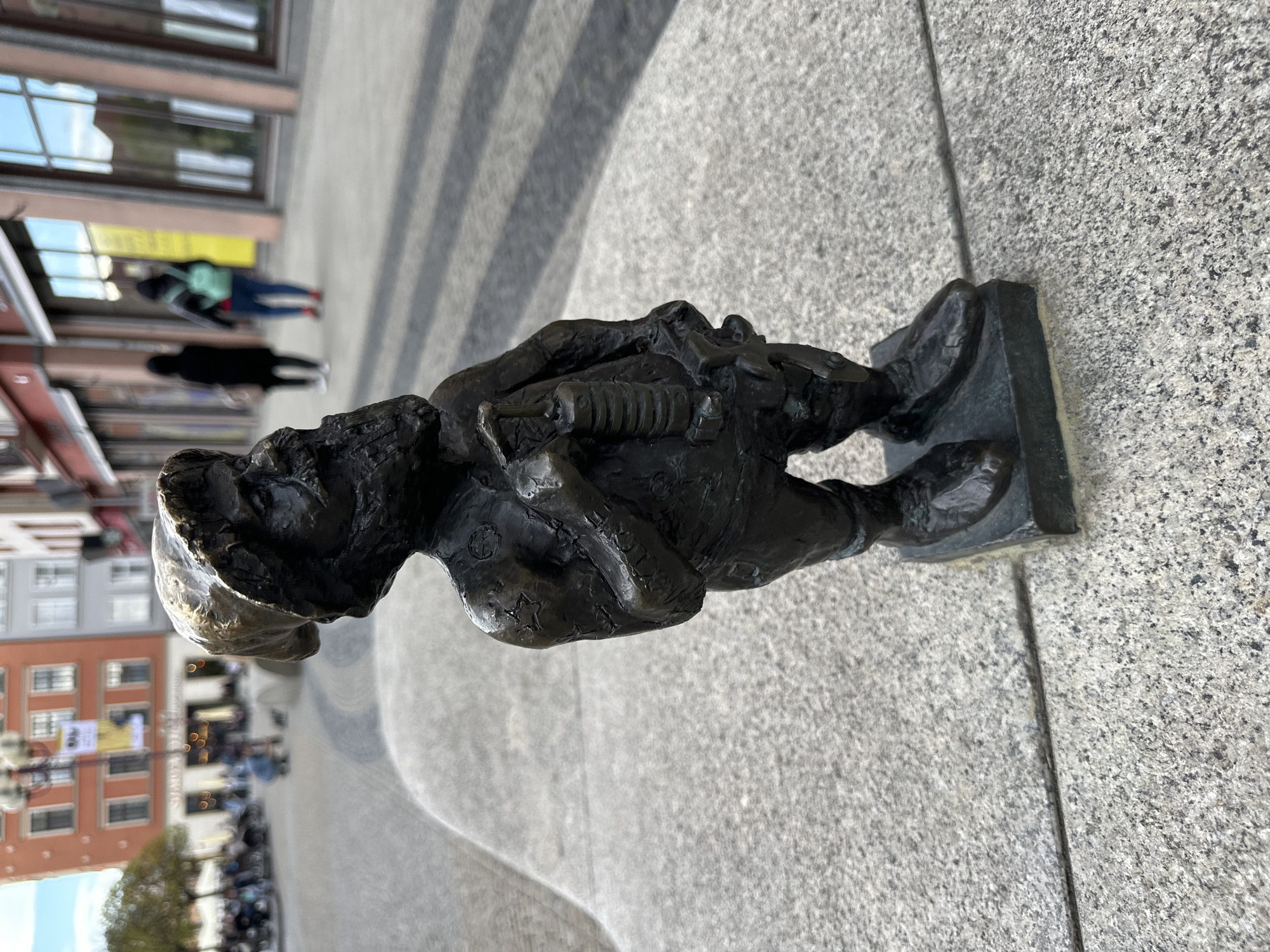 A bronze statue of a dwarf stands on a stone tile surface in Wrocław, Poland. The dwarf is wearing a hat and has a beard. He is holding a small object that resembles a musical instrument. In the background, there are buildings, people walking, and other urban features, suggesting a city square.